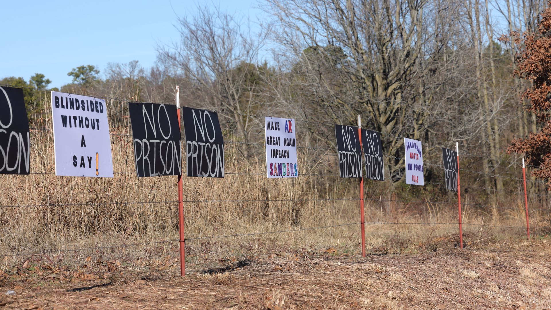 A series of signs protesting a prison are posted on a barbed wire fence. 