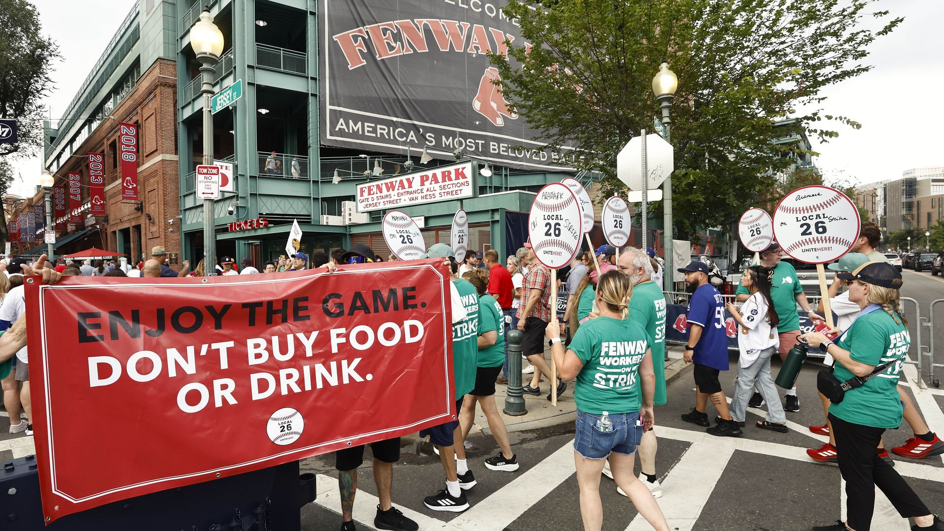 Protesters outside Fenway Park in green shirts holding baseball-shaped signs reading "Local 26 Strike" and a red banner saying "Enjoy the game. Don't buy food or drink."