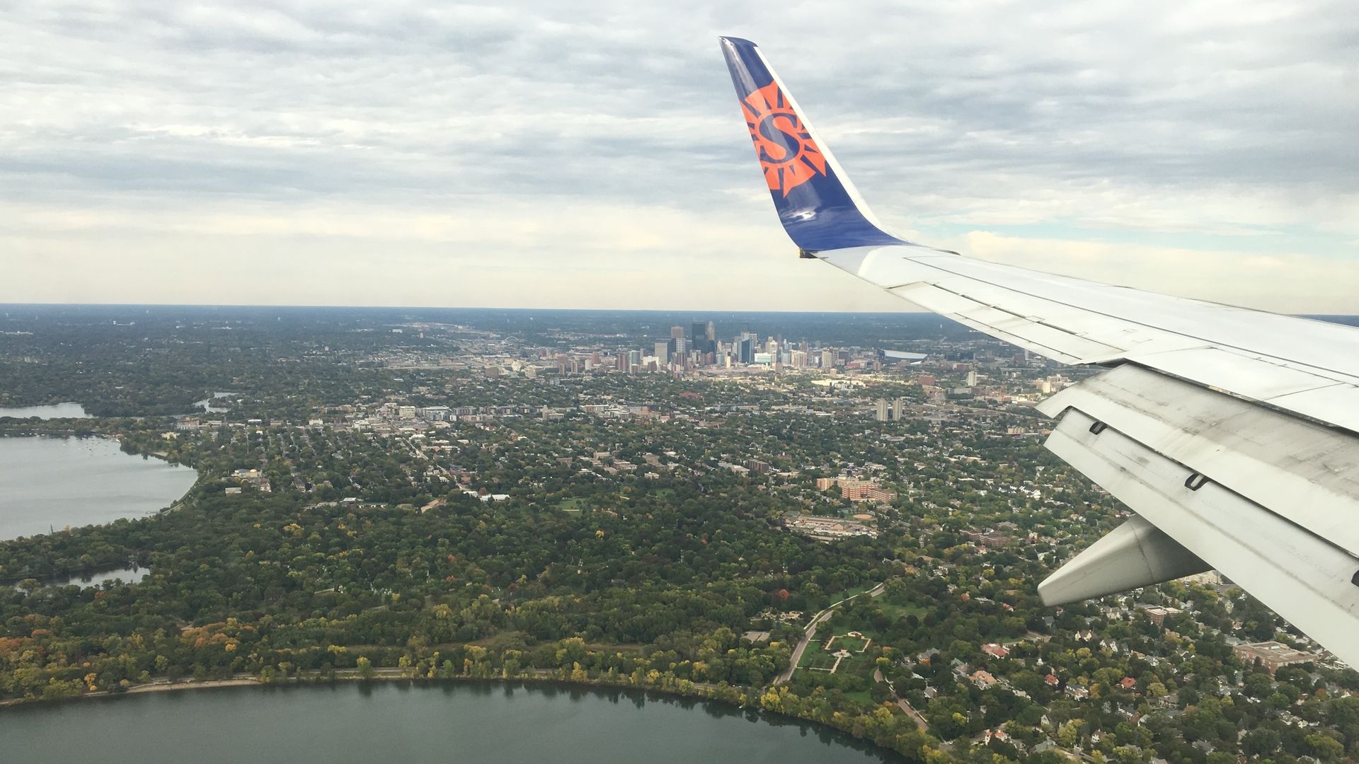 View from an airplane window: a white wing extends across the frame, blue tail with an orange sunburst logo, city skyline and river below, with green parks and a cloudy sky.