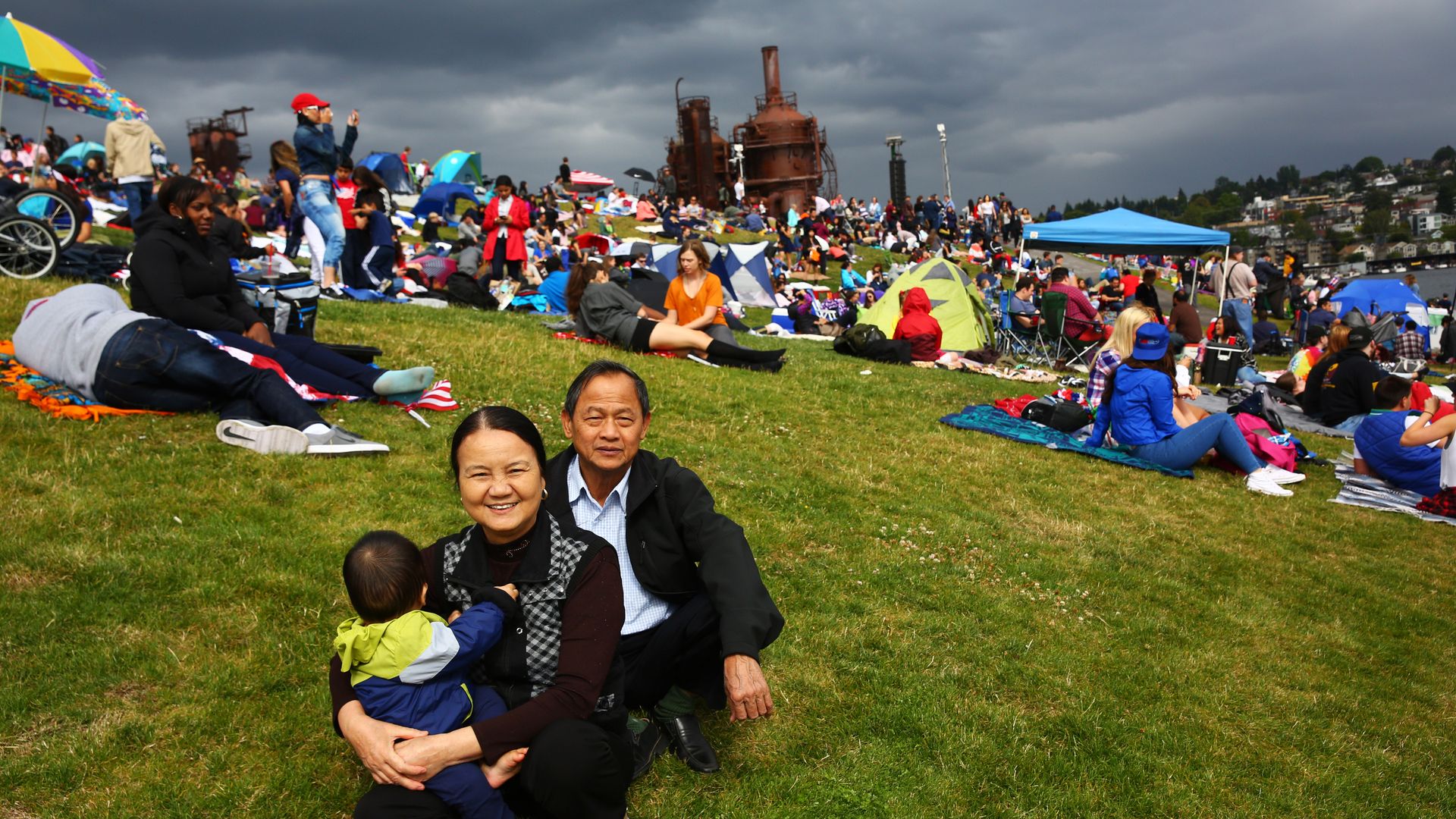 People gathered on the grass at Gas Works Park with the old gas works, which is brown, in the background.