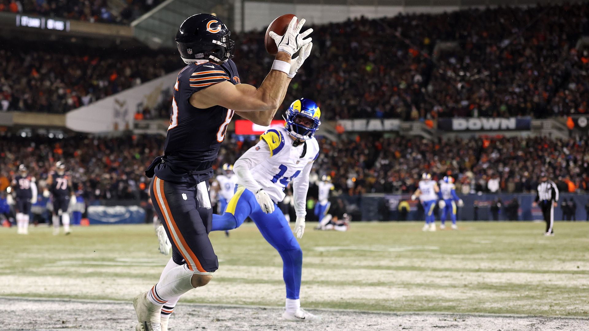 Chicago Bears player in navy and orange catches a football near the end zone while a Los Angeles Rams player in white and blue tries to defend during a night game.