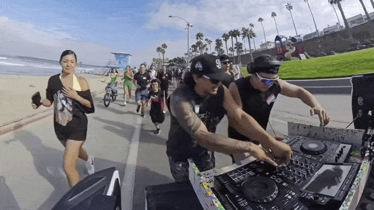 Two DJs mix on a colorful equipment deck at a sunny beach boardwalk while crowds stroll by. Palm trees line the walkway, the ocean sits to the left, and a lifeguard tower is visible in the distance.