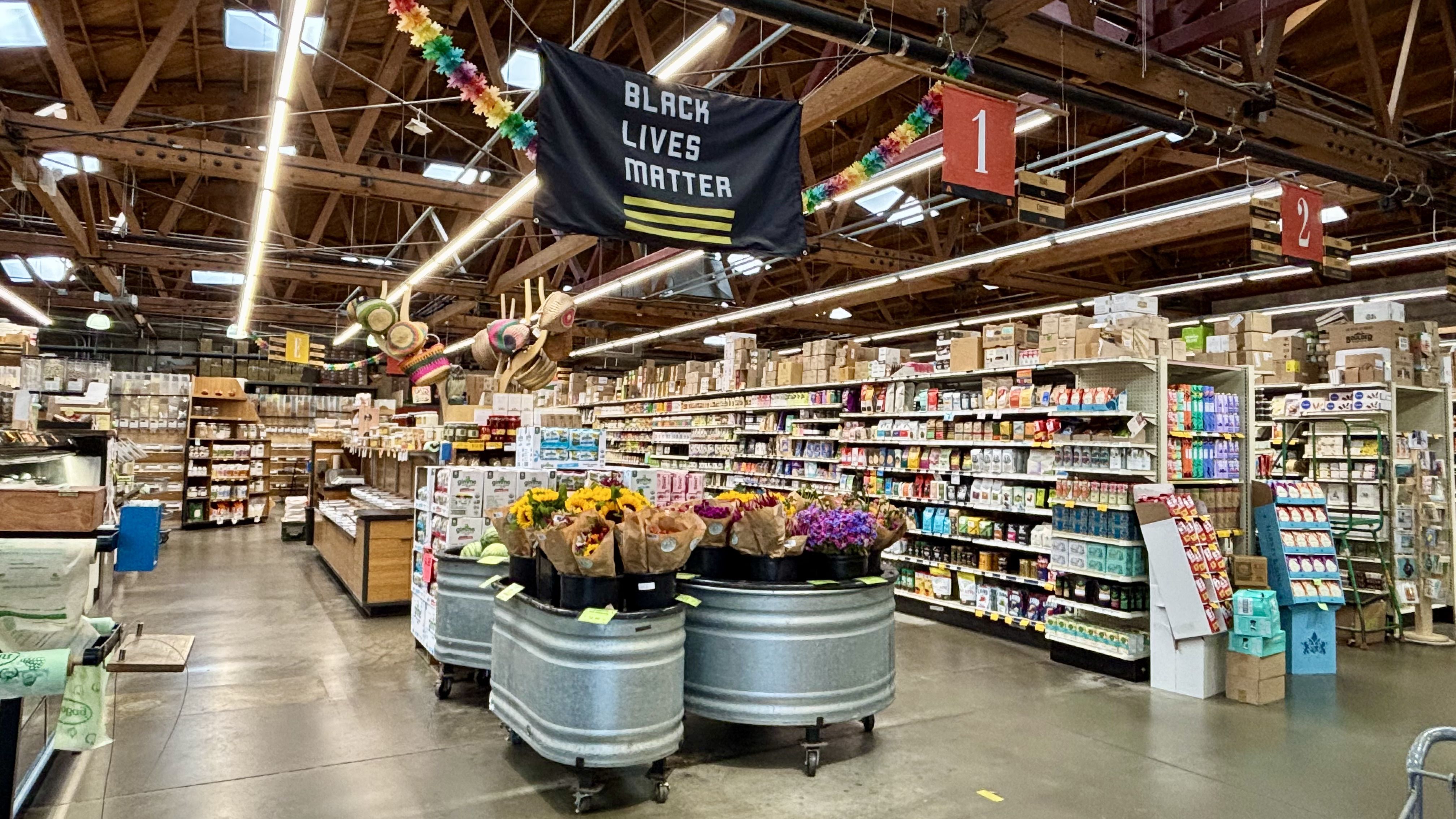 Interior of a grocery store with wooden ceiling beams, shelves stocked with groceries, flower bouquets in metal tubs, and a Black Lives Matter flag hanging from the ceiling.