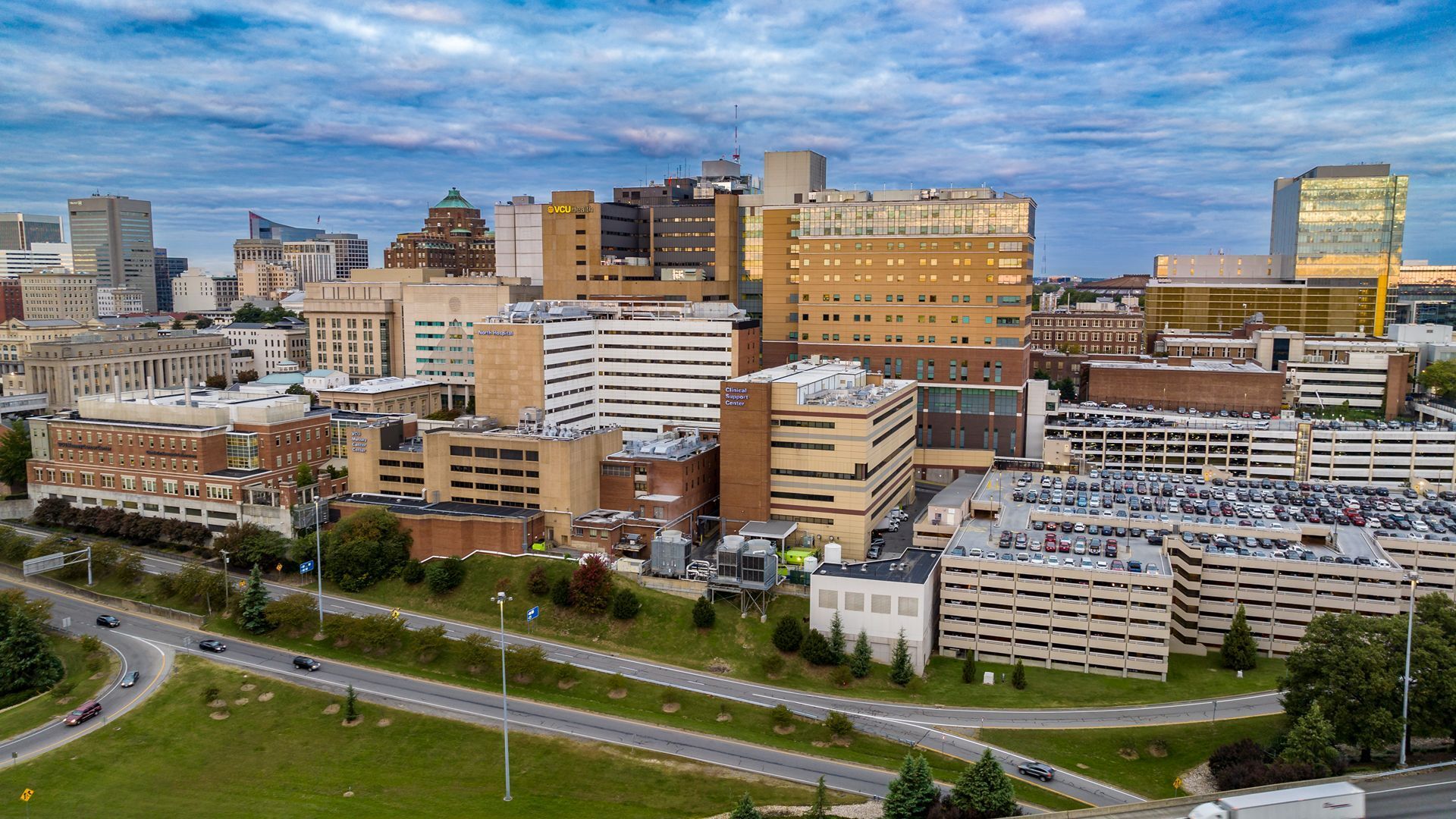 Aerial view of a downtown skyline with beige and glass office buildings, a hospital complex, and a large parking garage. Curving roads snake through green foreground under a blue, cloudy sky.