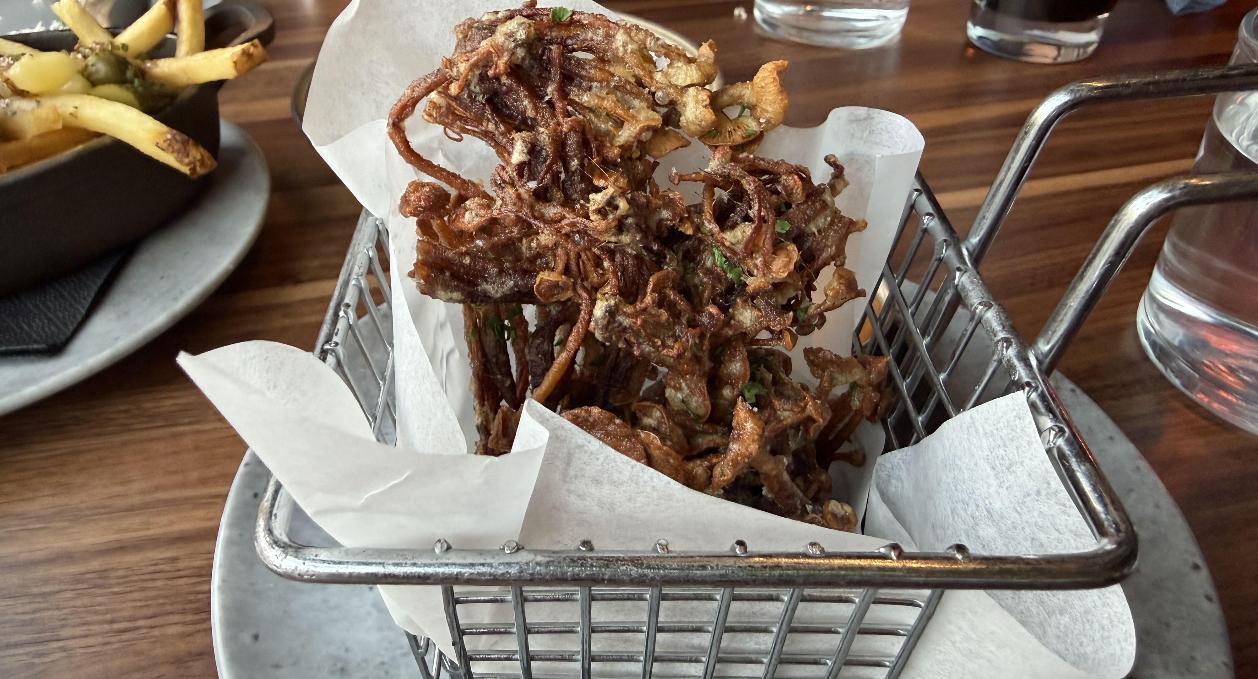 A metal basket lined with parchment holds a tall pile of crispy fried onion strings, golden brown and tangled, on a wooden table beside fries and water glasses.