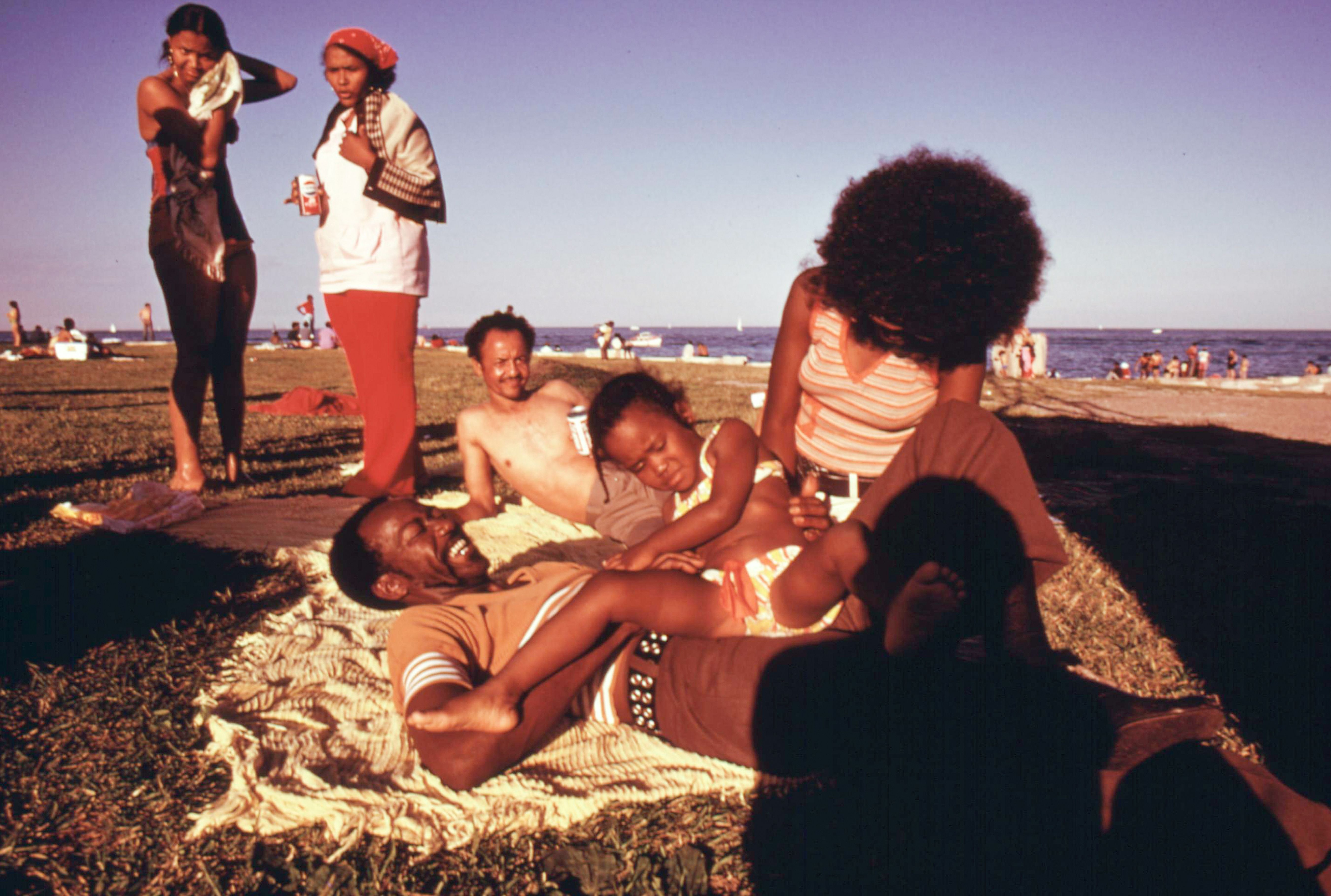 Beach scene: a woven blanket on the sand in the foreground, with a woman having a big curly afro wearing a striped top seated near the edge. Two women stand nearby; the blue sea and boats lie in the distance.