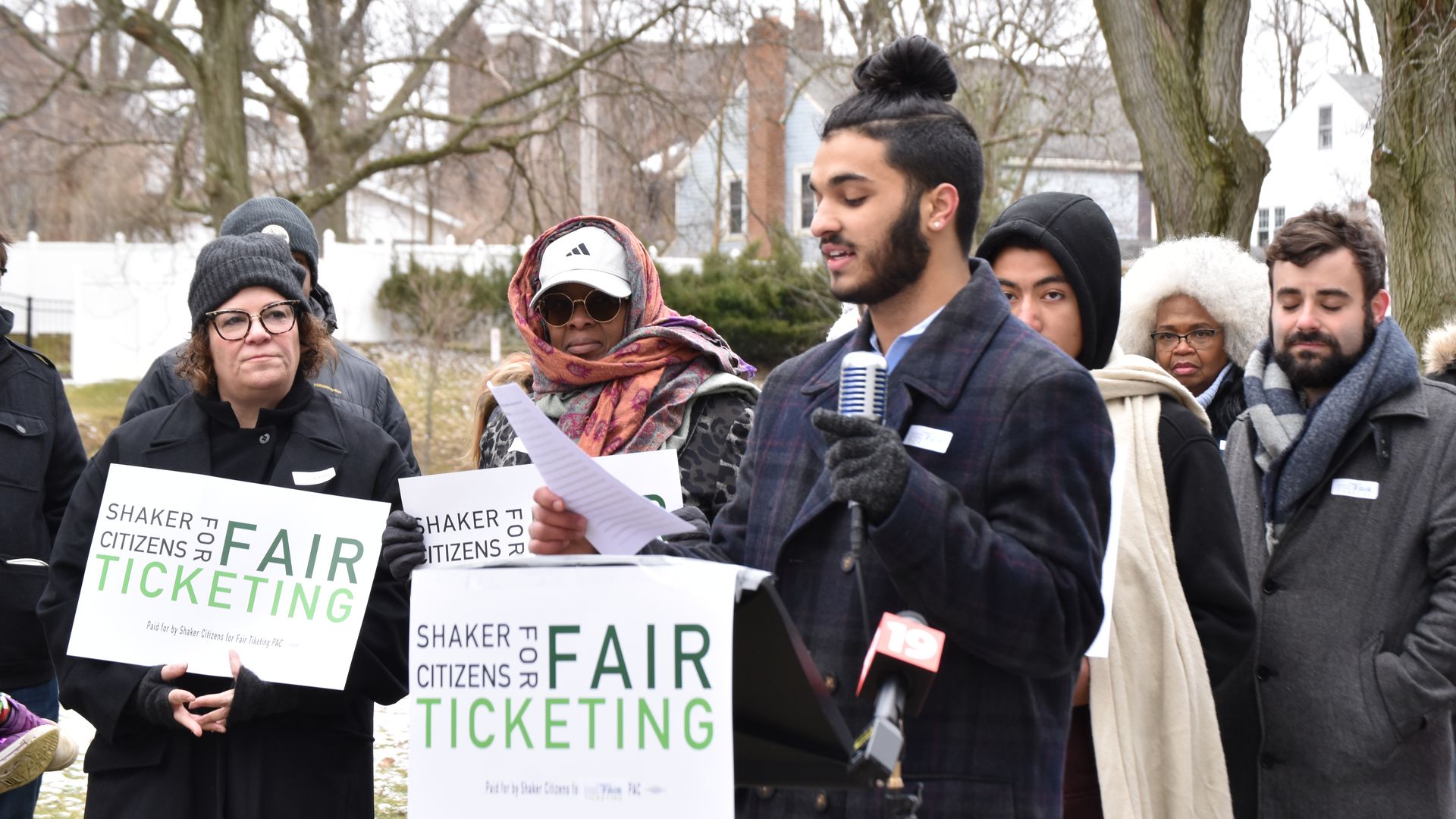 A young man speaks outdoors with a microphone, as people hold signs saying "Shaker Citizens for Fair Ticketing"