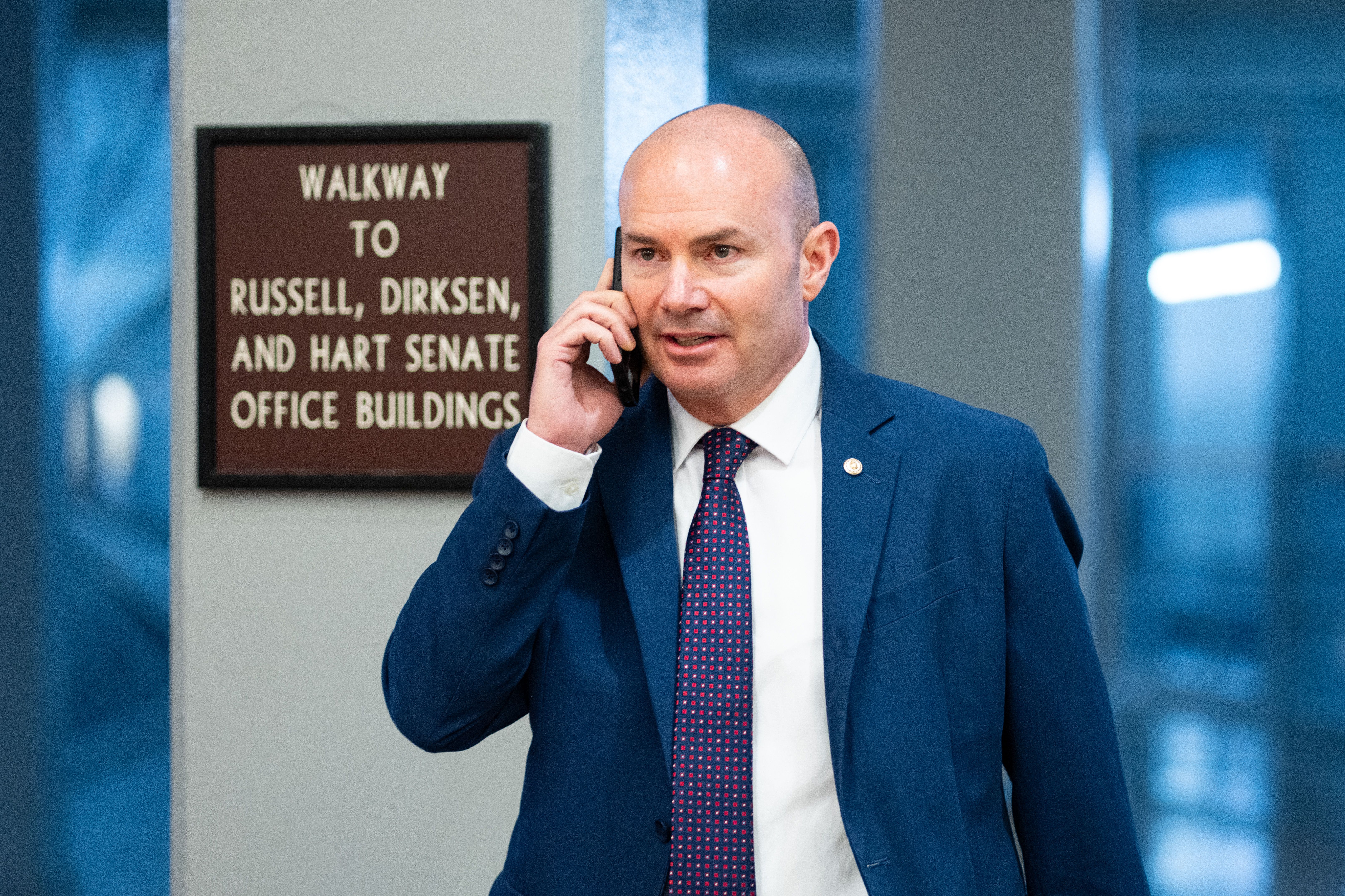 Man in a blue suit and patterned tie talking on a phone near a sign for walkway to Russell, Dirksen, and Hart Senate office buildings with a blue background.