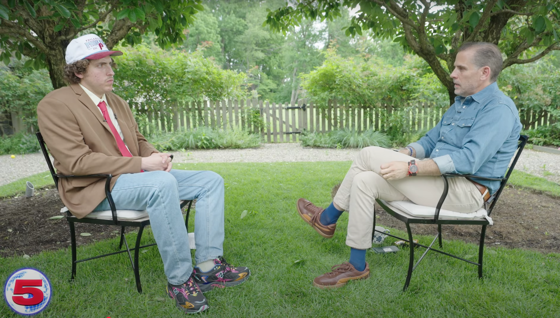 Two men seated on black metal chairs in a green garden with trees and a wooden fence in the background; one wears a brown blazer, red tie, and white cap, the other a blue denim shirt and beige pants.