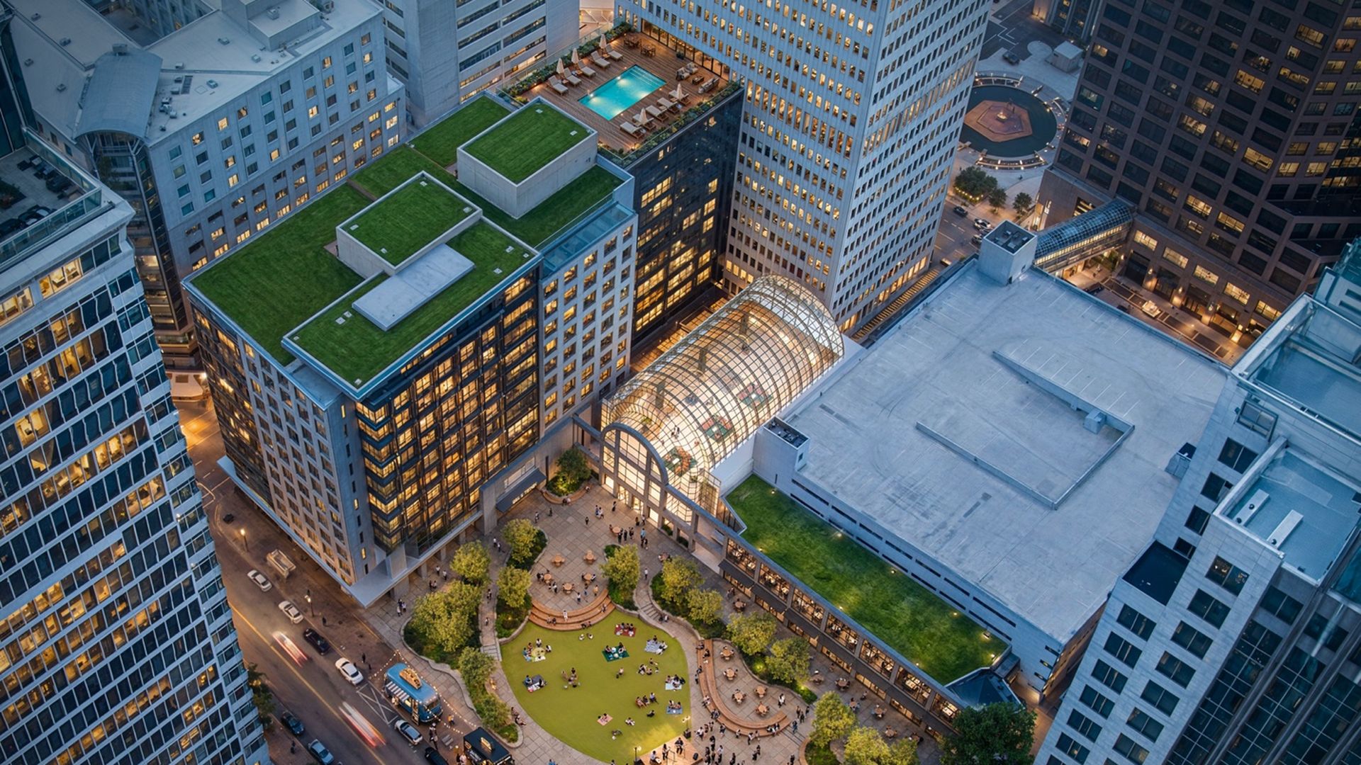 Aerial view of a city plaza surrounded by tall glass skyscrapers. A glass-domed arcade anchors a lively courtyard with green rooftop gardens, trees, and people strolling.