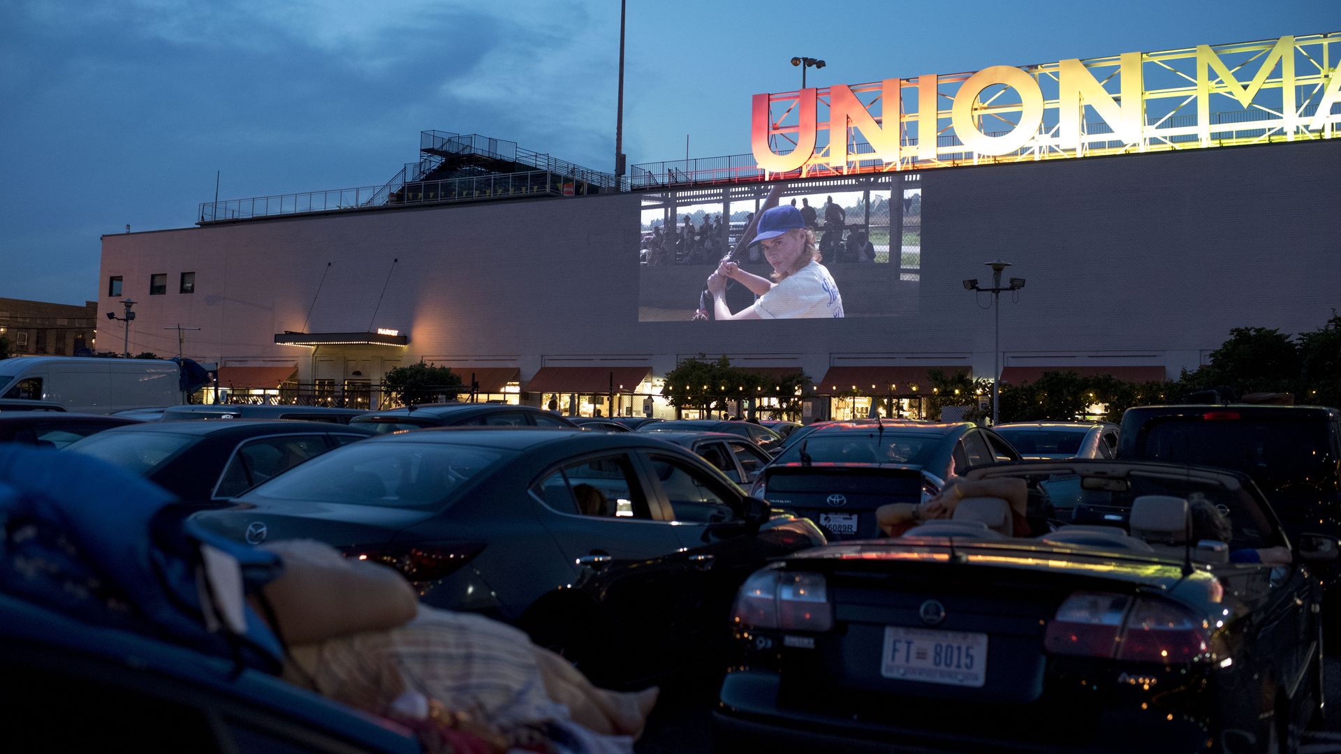 In this image, a girl holding a baseball bat is projected on the side of a building in front of cars