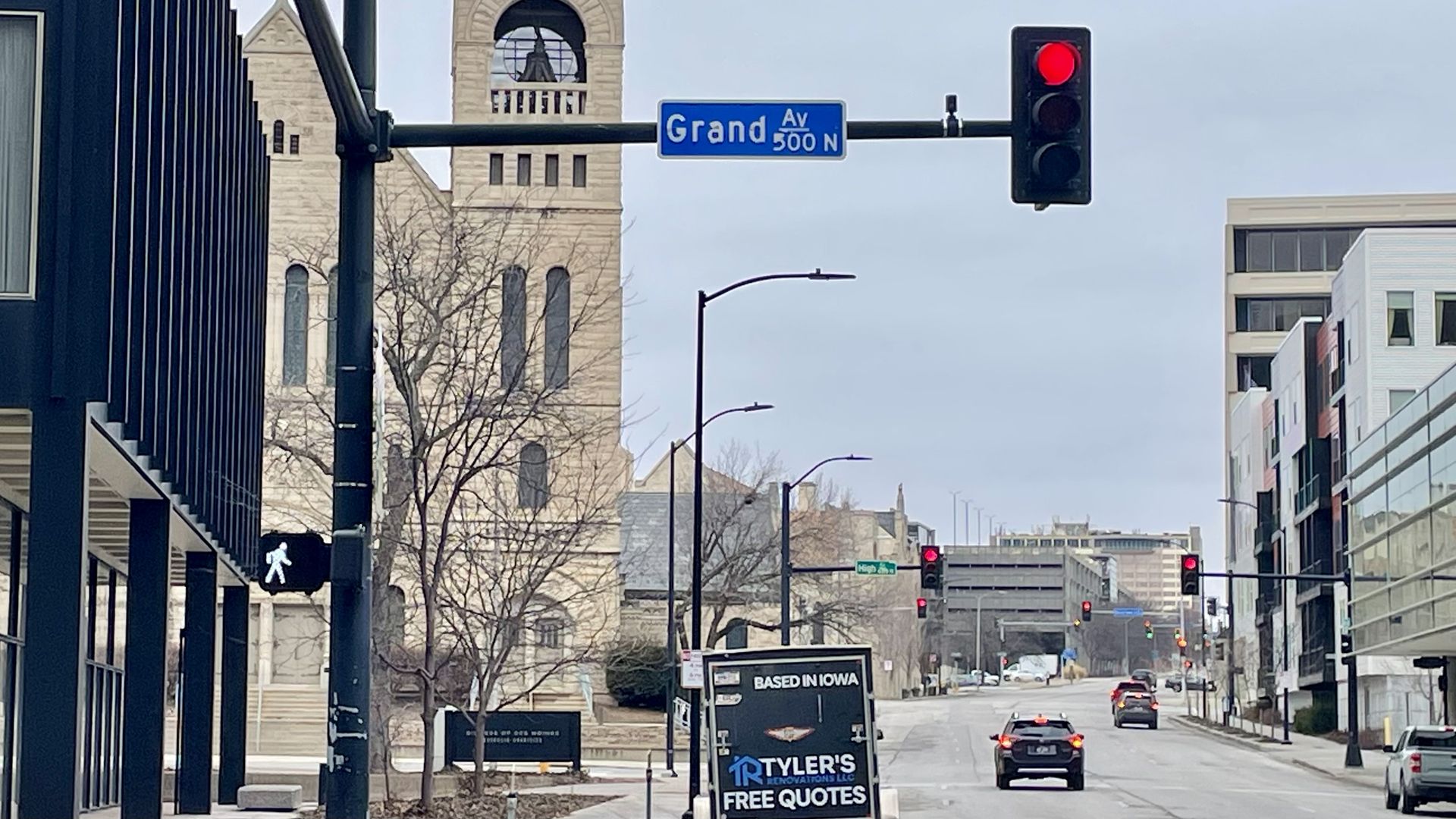 Urban street scene at Grand Avenue with traffic lights showing red for vehicles and white for pedestrians. Buildings and leafless trees line the road under a gray sky.