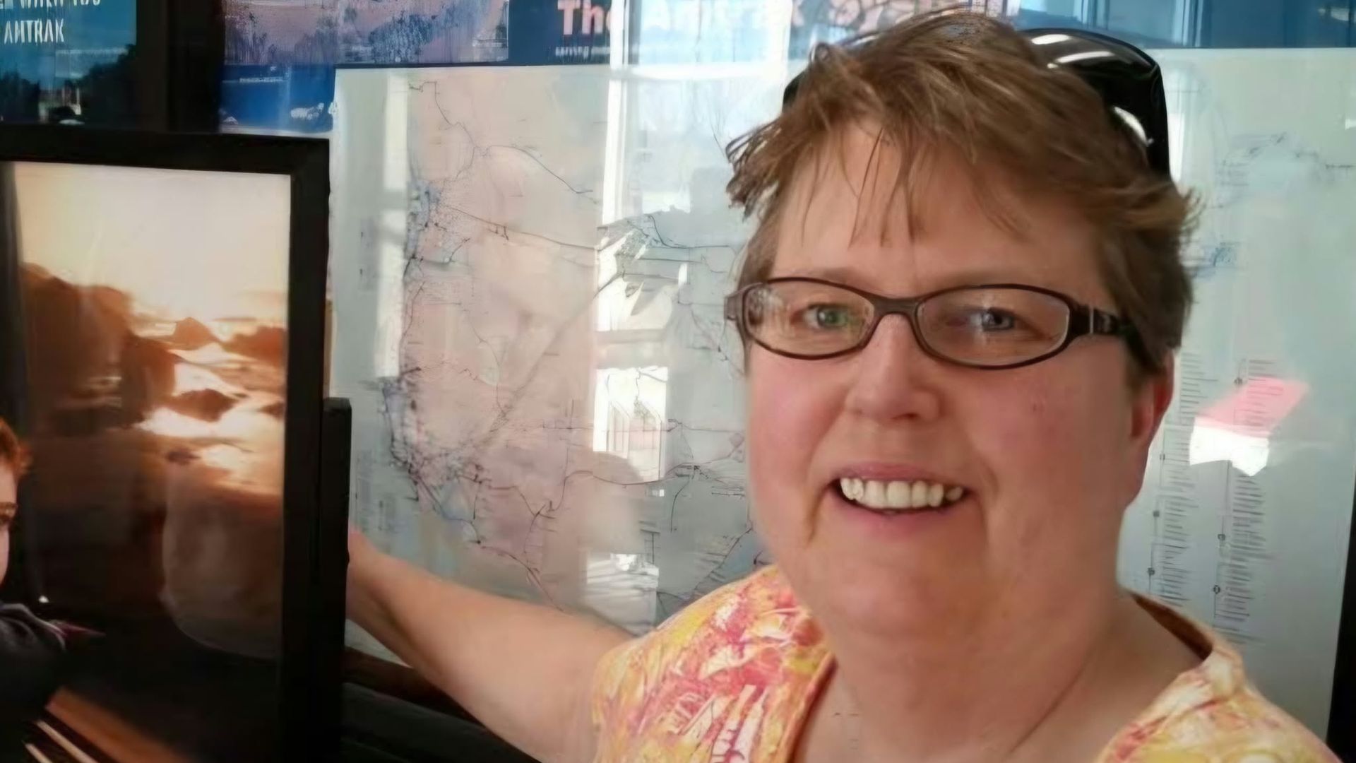 Smiling person with short brown hair and glasses wearing a bright, patterned top, posing in front of a glass wall covered with maps and travel posters; reflections show Arrivals and Departures.