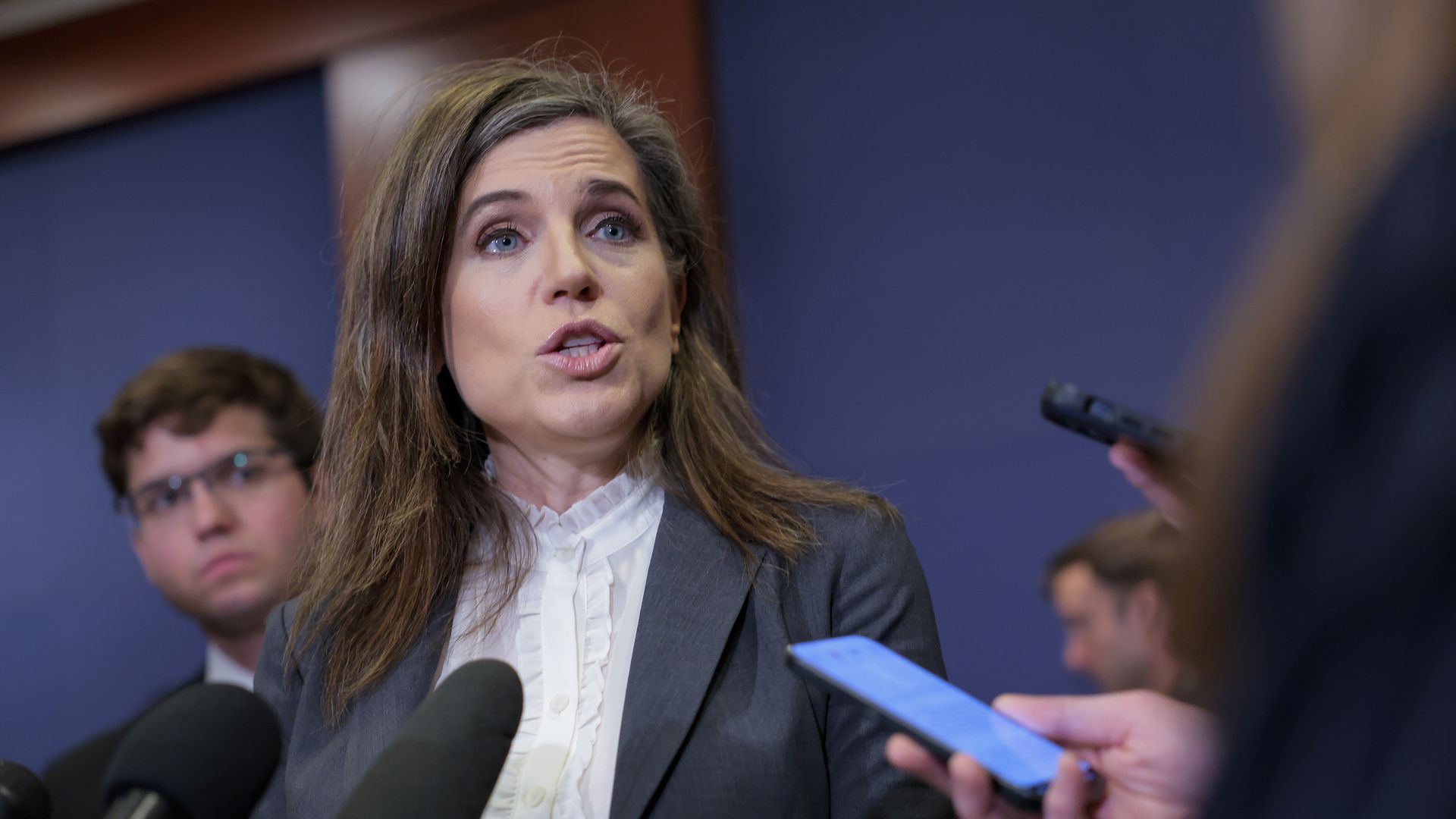 Spokeswoman in a gray blazer and white ruffled blouse speaks to reporters, with multiple microphones in front; a man with glasses and a person holding a blue phone appear in the background.
