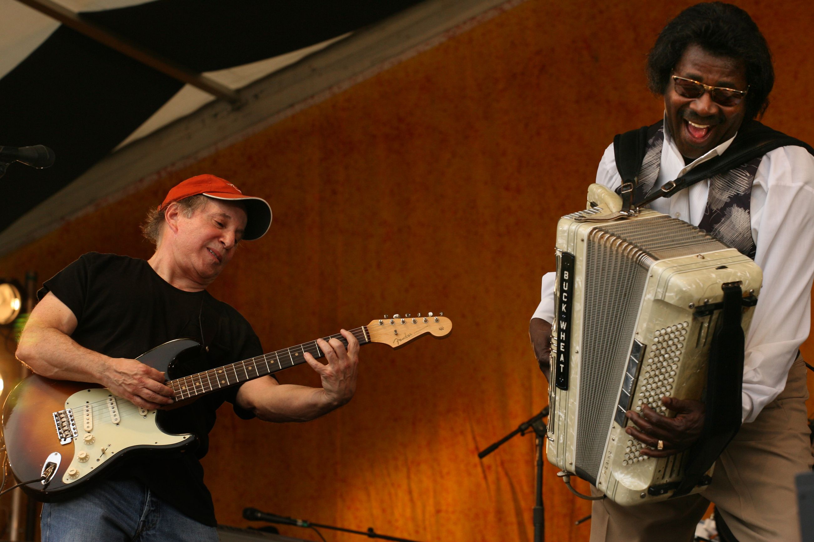 Two musicians on stage playing warmly; one in a red cap with a black Fender electric guitar, the other smiling widely, wearing sunglasses and playing a cream-colored accordion labeled "Buck Wheat".