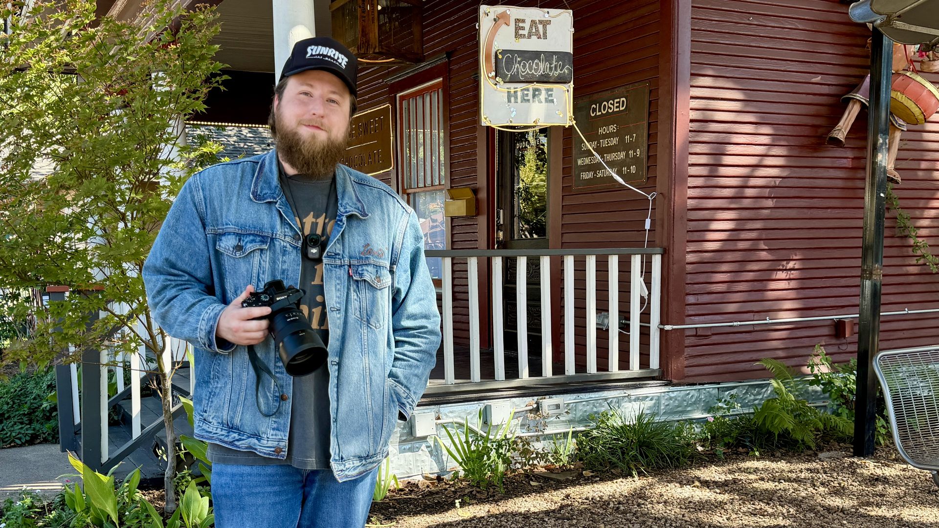 Bearded man in a blue denim jacket and black cap holds a camera outside a brown storefront with signs reading EAT and Chocolate Here, surrounded by green plants and a white railing.