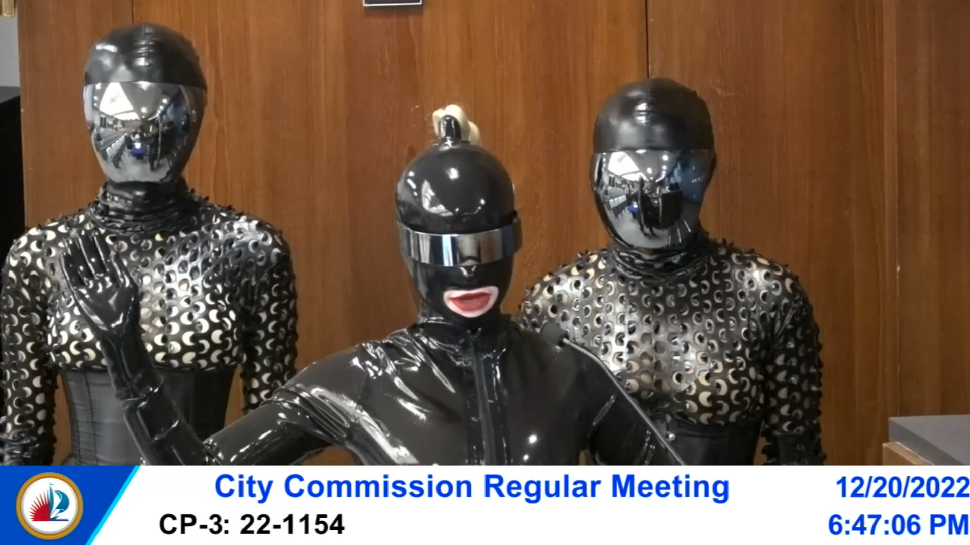 Three women in tight vinyl catsuits and masks stand in front of a wood-paneled wall 