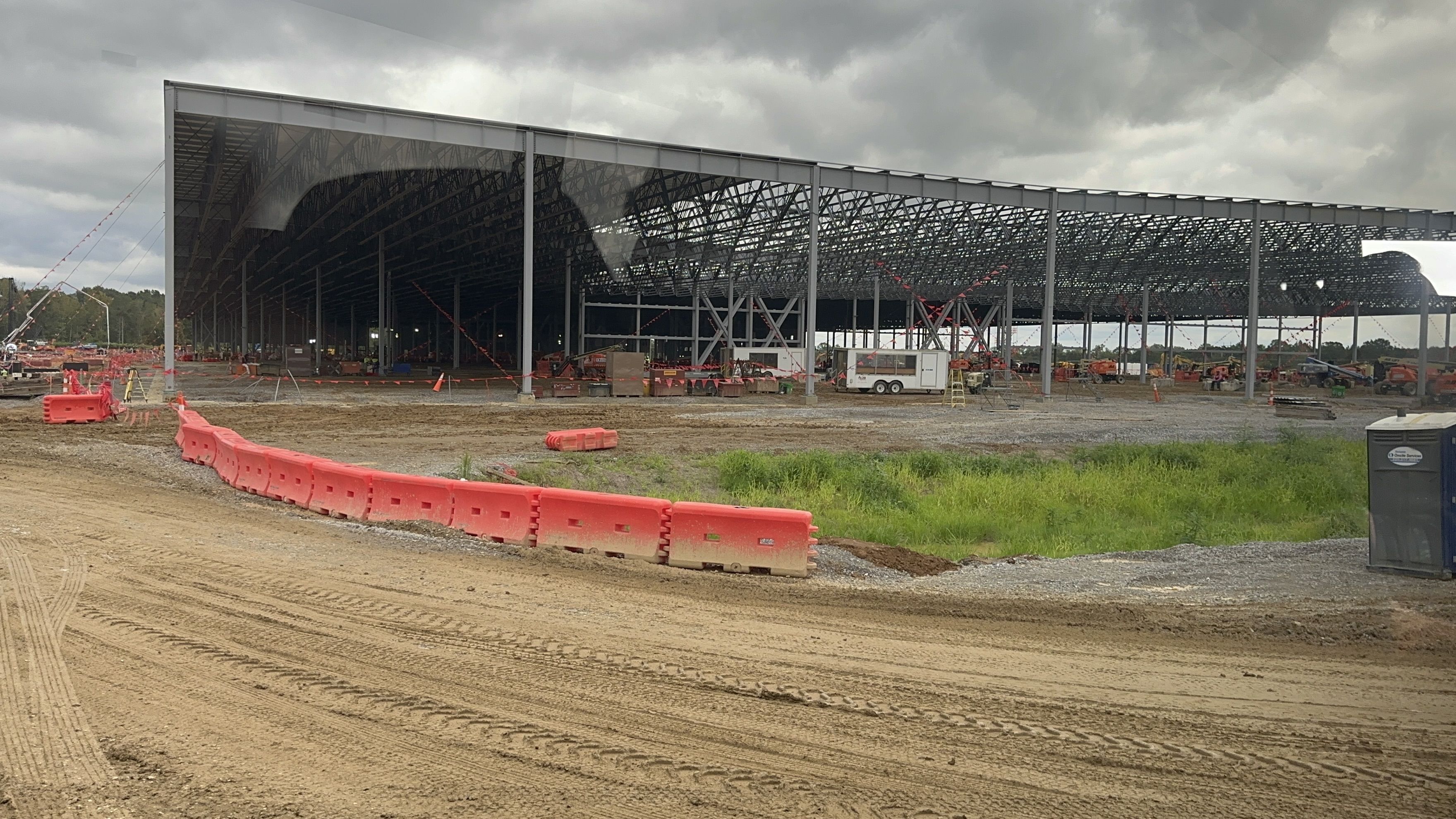 Under construction, a large steel-framed warehouse with an open truss roof, red safety barriers lining muddy ground, construction equipment scattered around, and a cloudy sky.