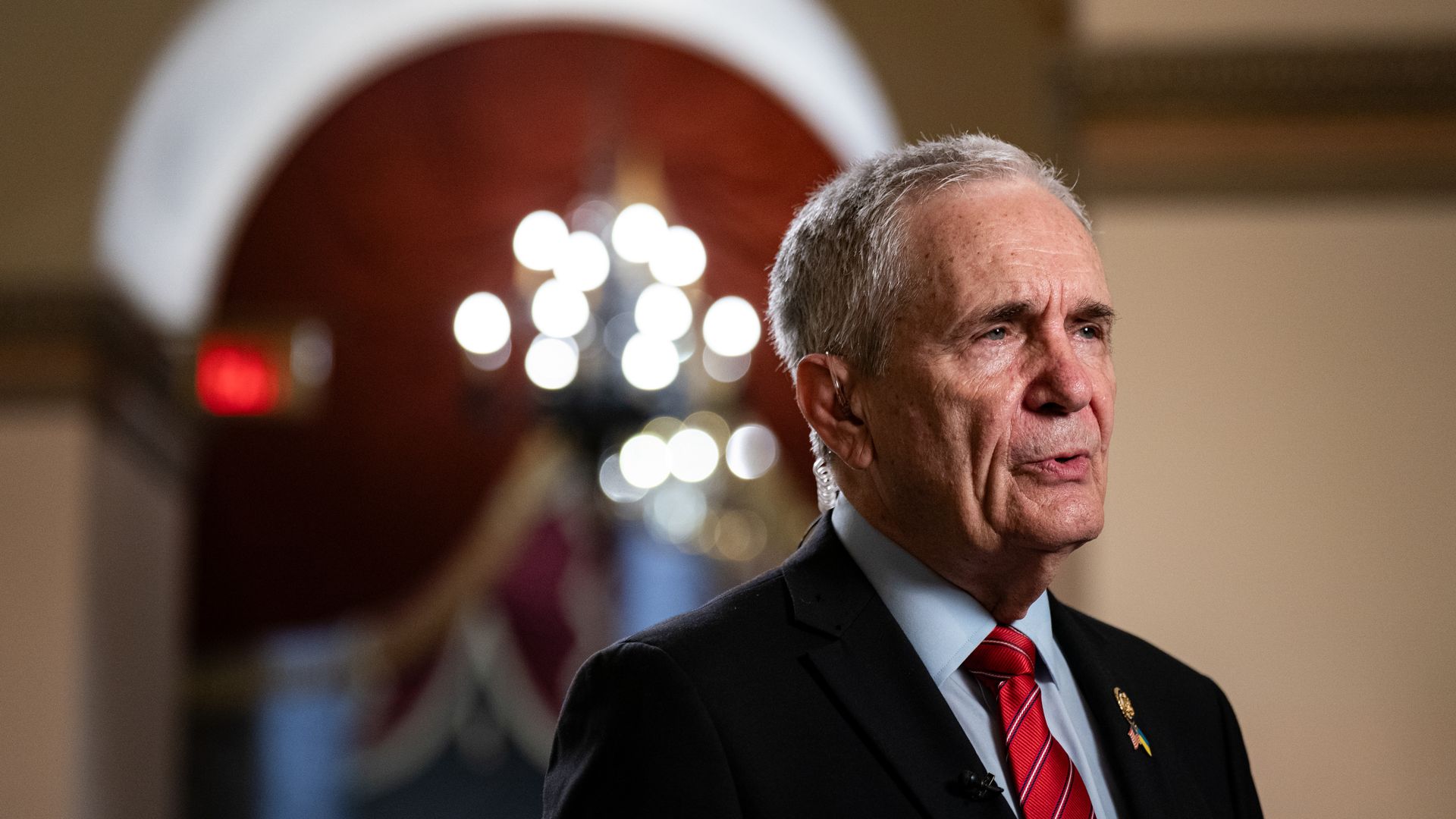 Older man with gray hair wearing a black suit, light blue shirt, and red striped tie speaking indoors with blurred chandelier and red archway in background.