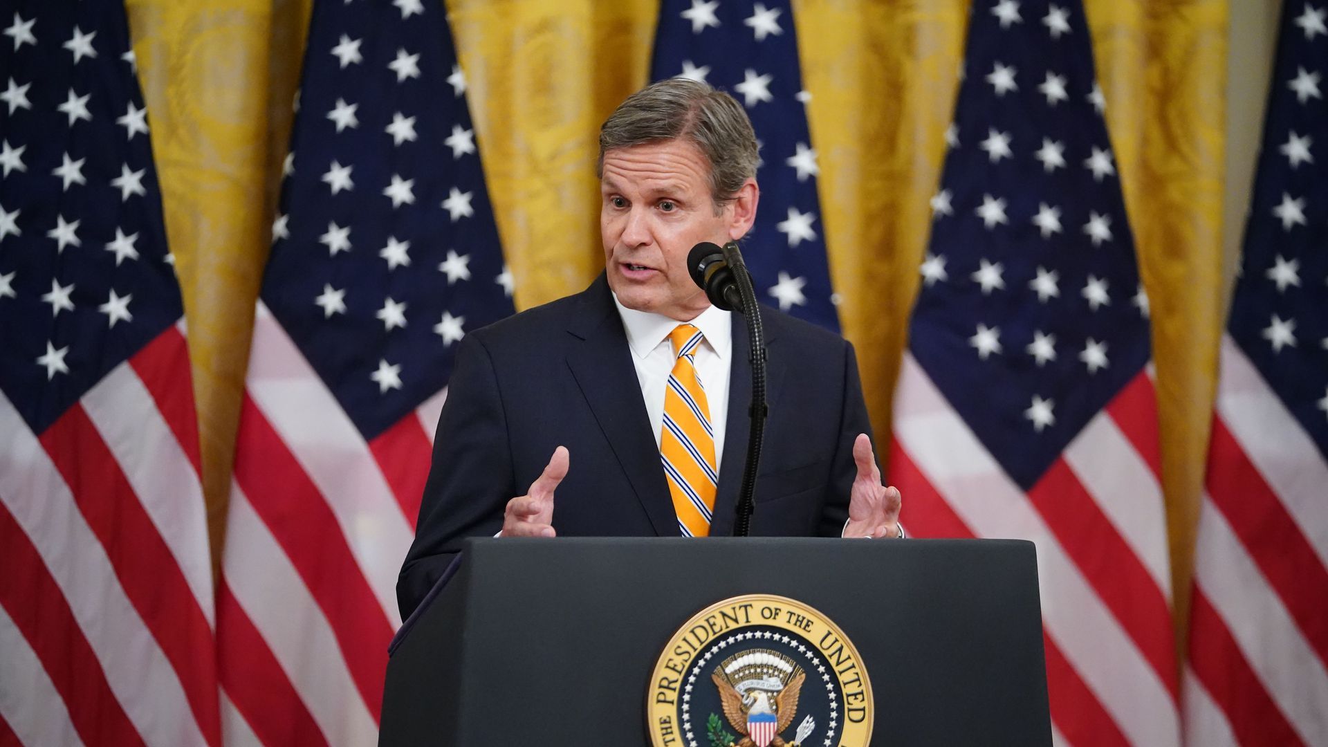 Tennessee governor Bill Lee in an orange tie and blue suit behind a lectern with American flags behind him