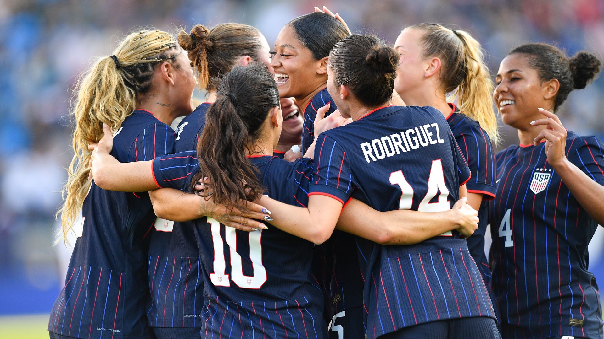 Group of women's soccer players in dark blue pinstriped uniforms huddle in celebration on the field, arms around each other, smiling; one jersey reads RODRIGUEZ with a USA crest.