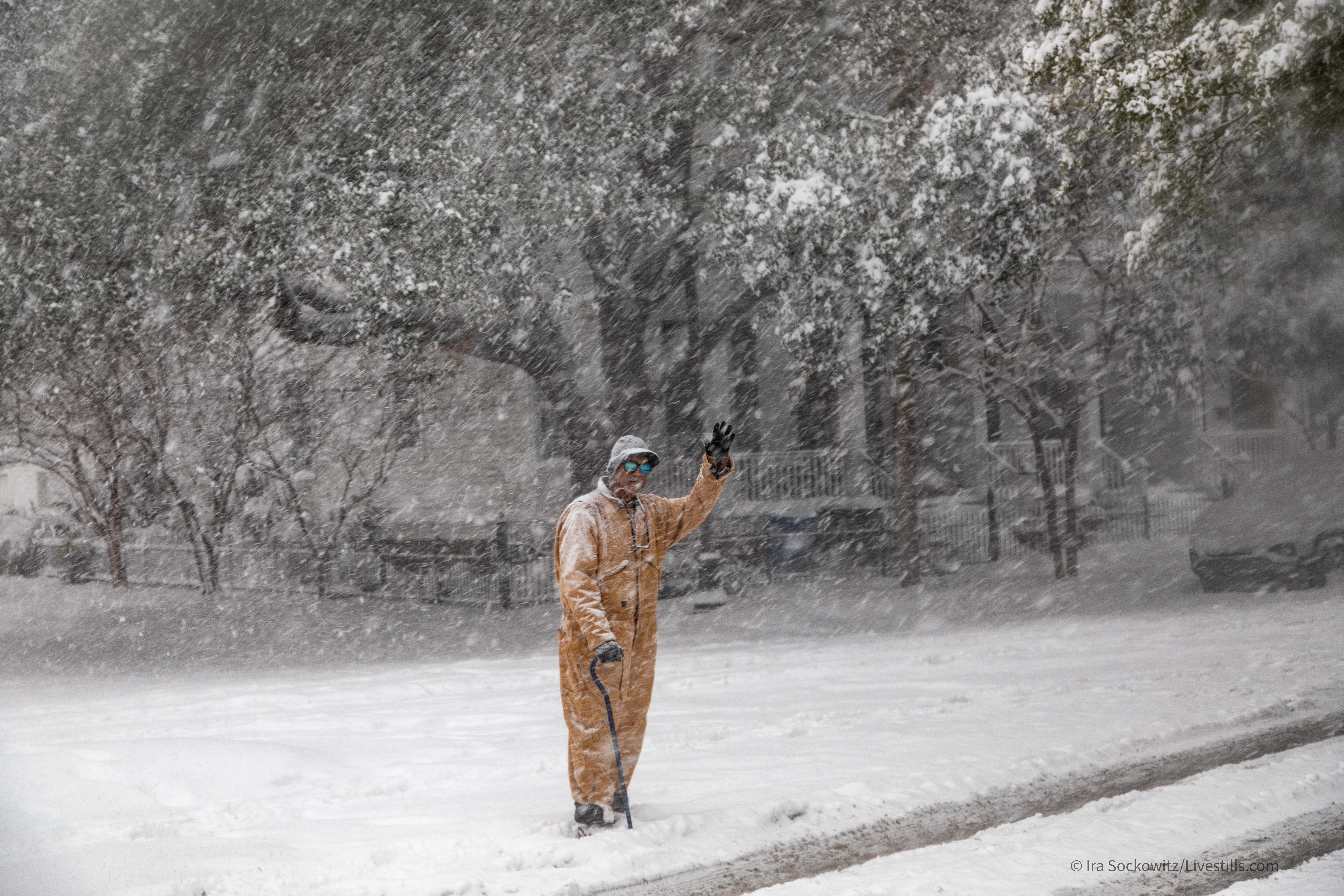 A man stands on a street's edge and waves in the snow.