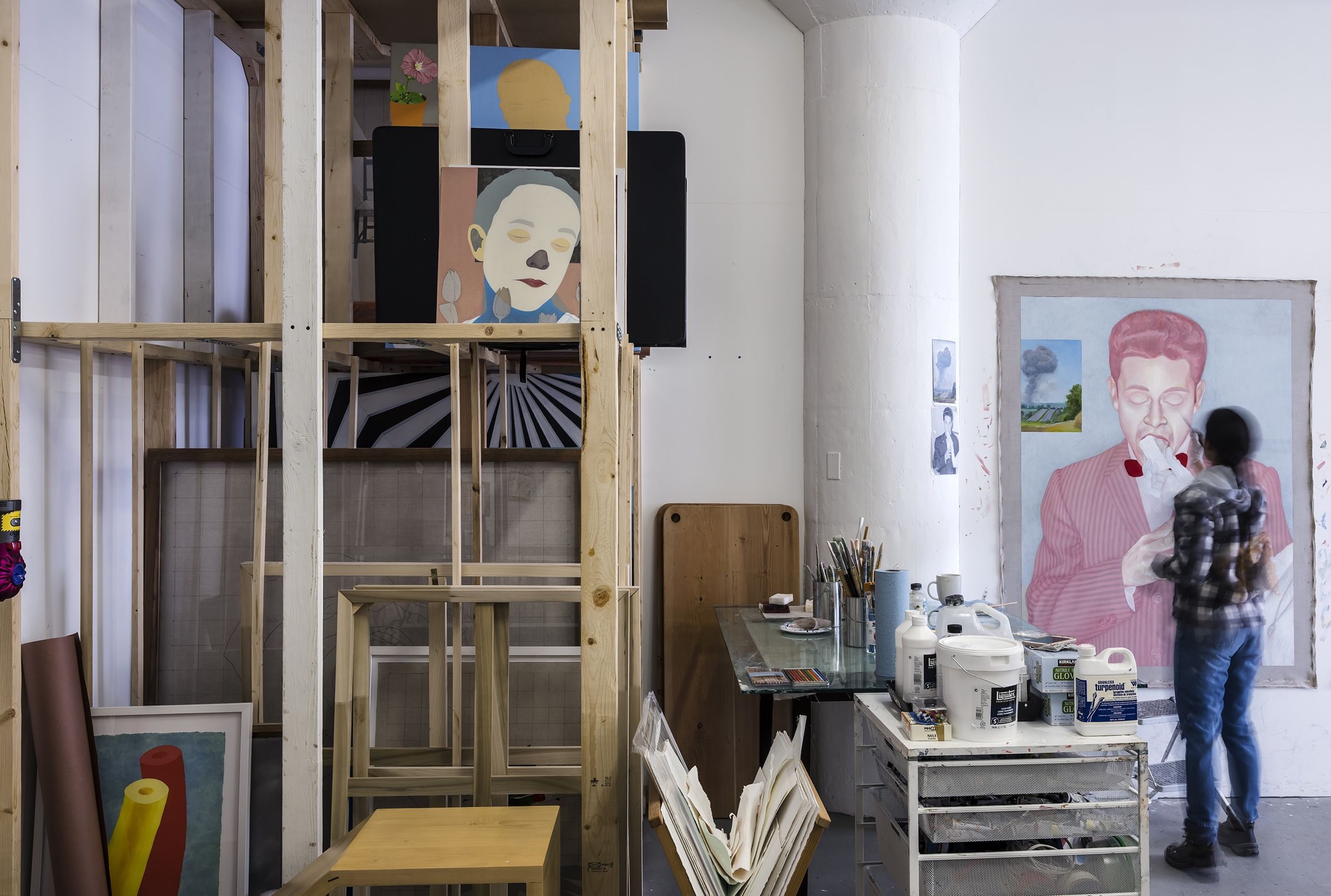 Plywood with some paintings leaning against it with a blurry person working on a painting of a man that's all in pink on the wall. Separated by a table with painting supplies.