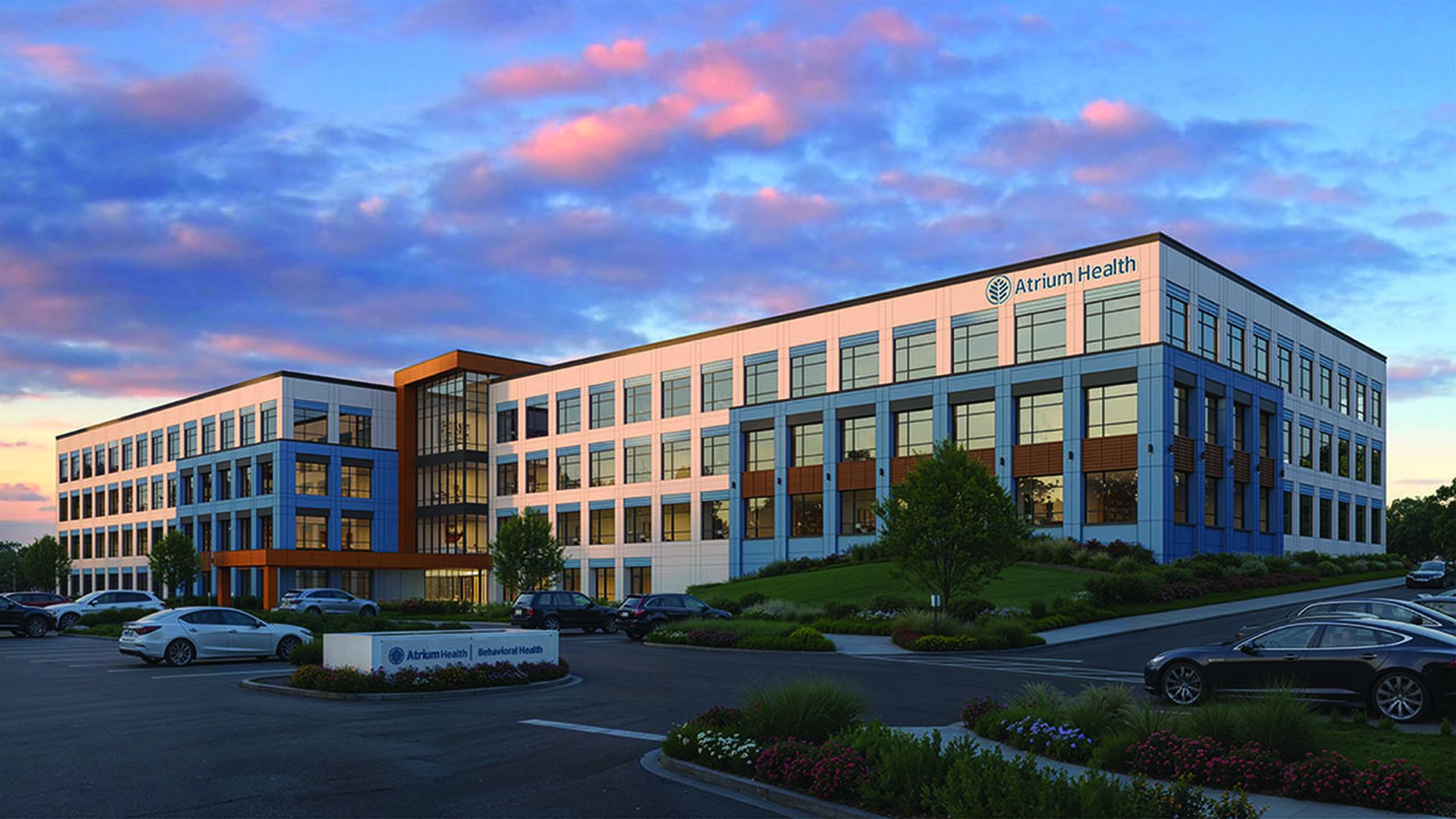 A modern blue-and-white office building with a glass central atrium and the "Atrium Health" sign. A parking lot with several cars and manicured landscaping sits in front at sunset.