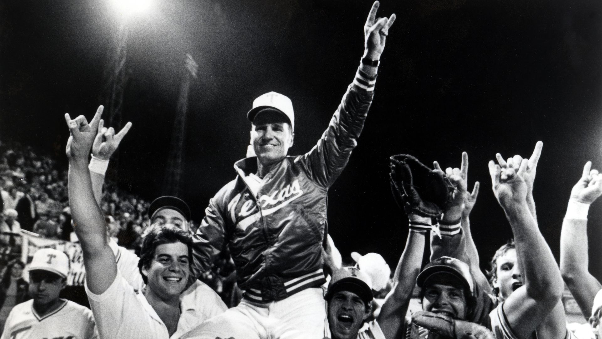 Black-and-white photo of Texas baseball players celebrating on the field; a man is lifted by teammates, raising his arm in victory as fans cheer in the stands.