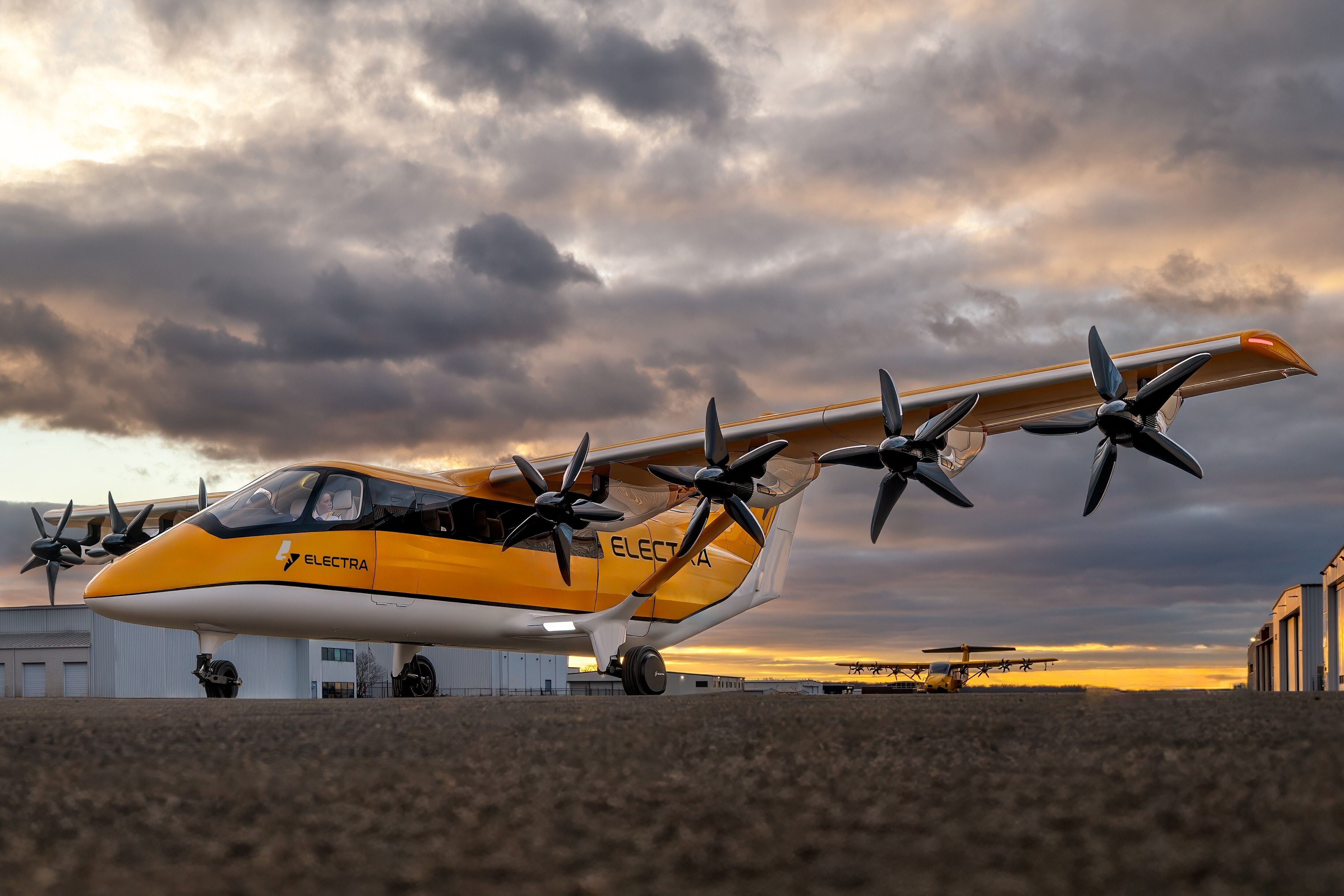 Bright yellow Electra seaplane with multiple propellers along its wing sits on a tarmac beneath a dramatic cloudy sunset; hangar buildings line the background.