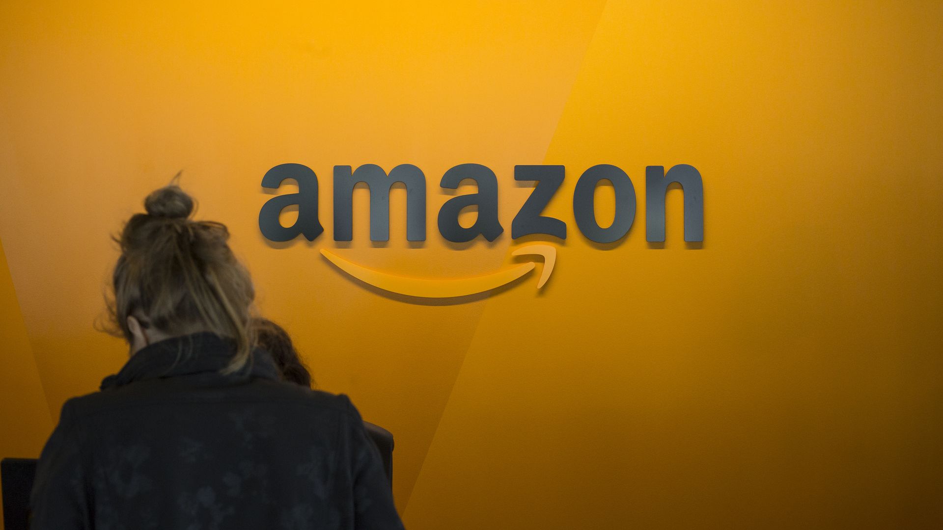 A woman walks in front of an orange wall with the Amazon.com logo on it