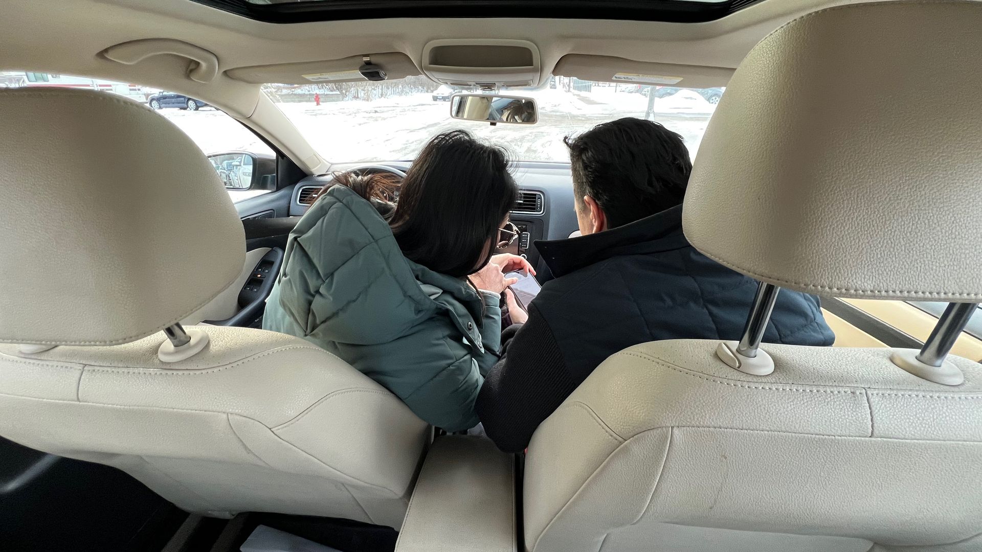 Rear view of two people sitting in front seats of a beige car, one in light blue jacket and the other in dark jacket, looking at a phone, with a snowy street visible through the windshield.