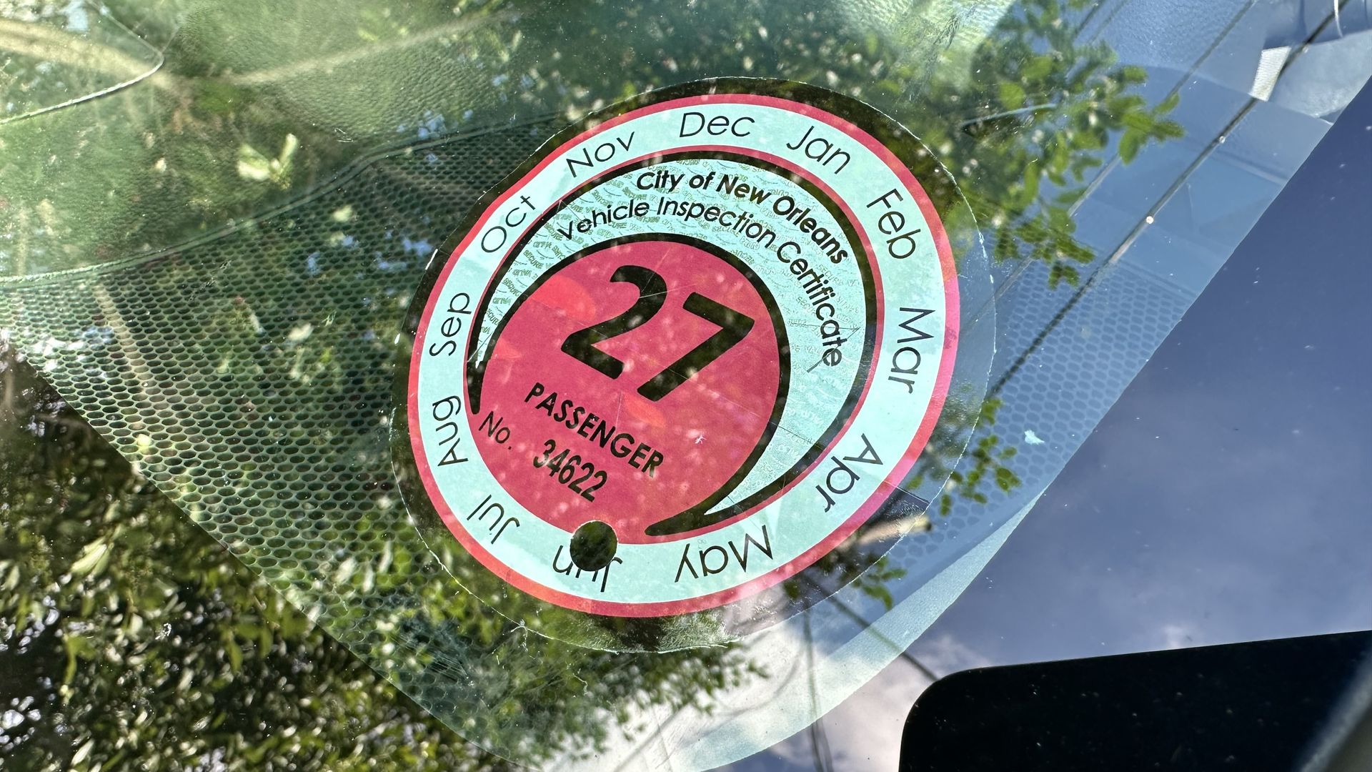 Close-up of a circular inspection sticker on a car windshield. Teal ring with month names surrounds a red center that reads City of New Orleans Vehicle Inspection Center.