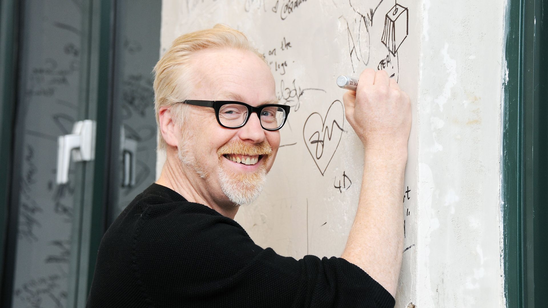 Photo of Adam Savage smiling at the camera as he signs a wall