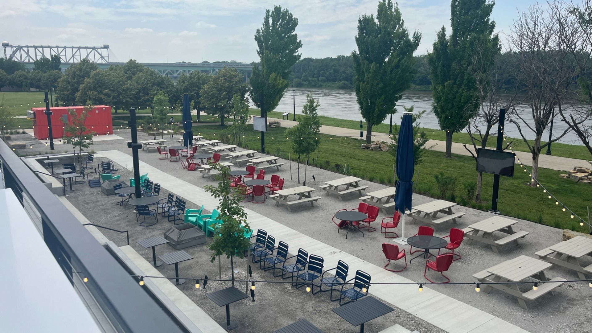 Photo from balcony looking down onto a beer garden lined with trees and full of red, teal, and blue colored chairs and tables.