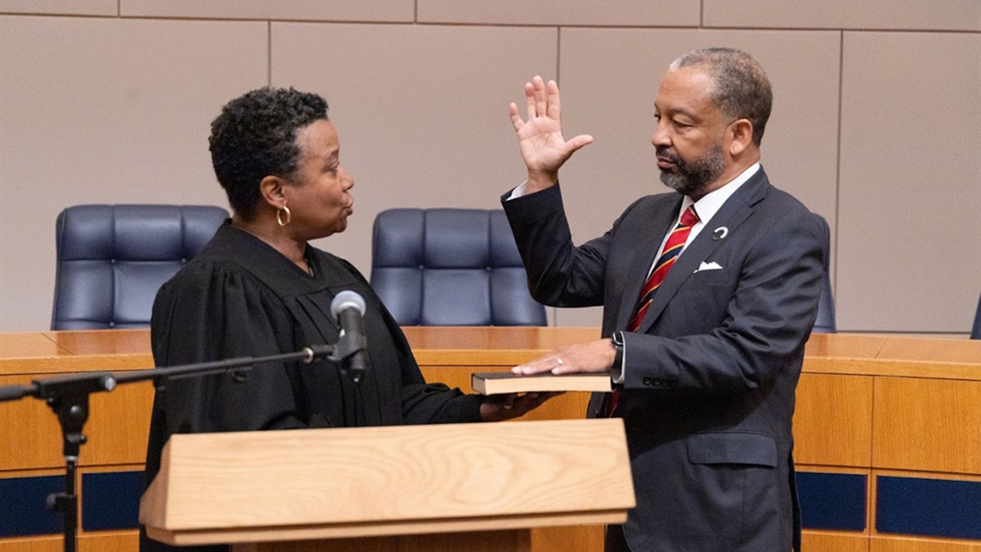 Man in a dark suit and red striped tie raises his right hand as a judge in a black robe administers an oath in a courtroom, with a book resting on the wooden podium.