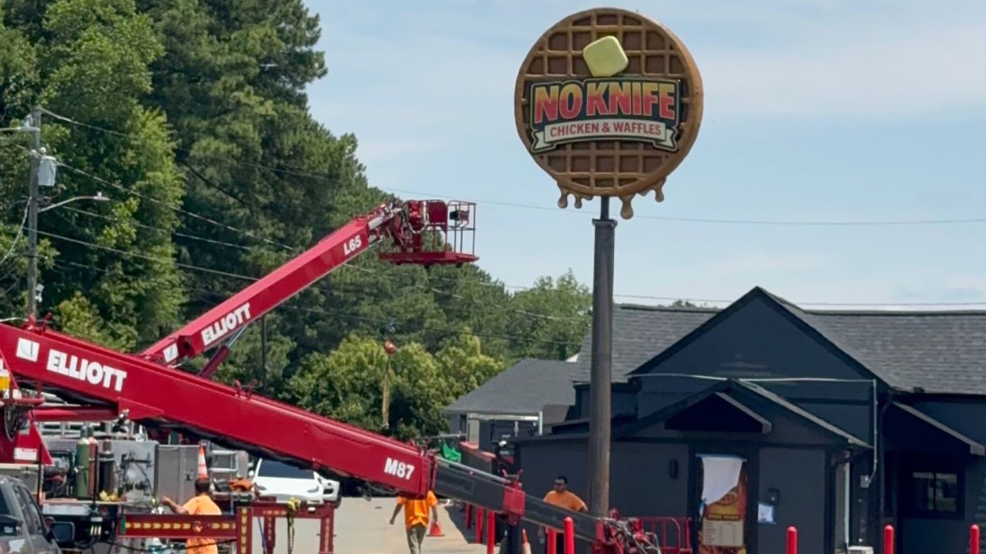 Clear blue sky above a street with construction workers in orange vests, orange cones, and a tall round waffle-sign reading "NO KNIFE CHICKEN & WAFFLES" beside a red crane.