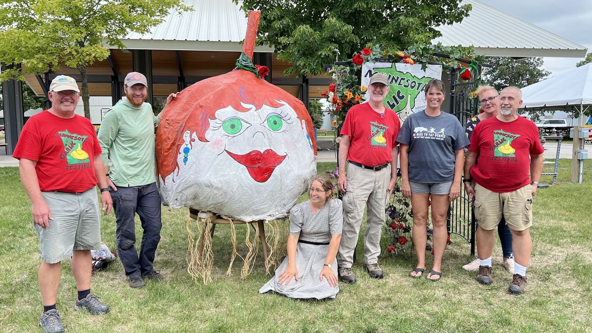 Group of seven people posing outdoors on grass next to a large red garlic sculpture with a face; three men wear red "Minnesota Garlic Festival" shirts, one woman sits by the sculpture.