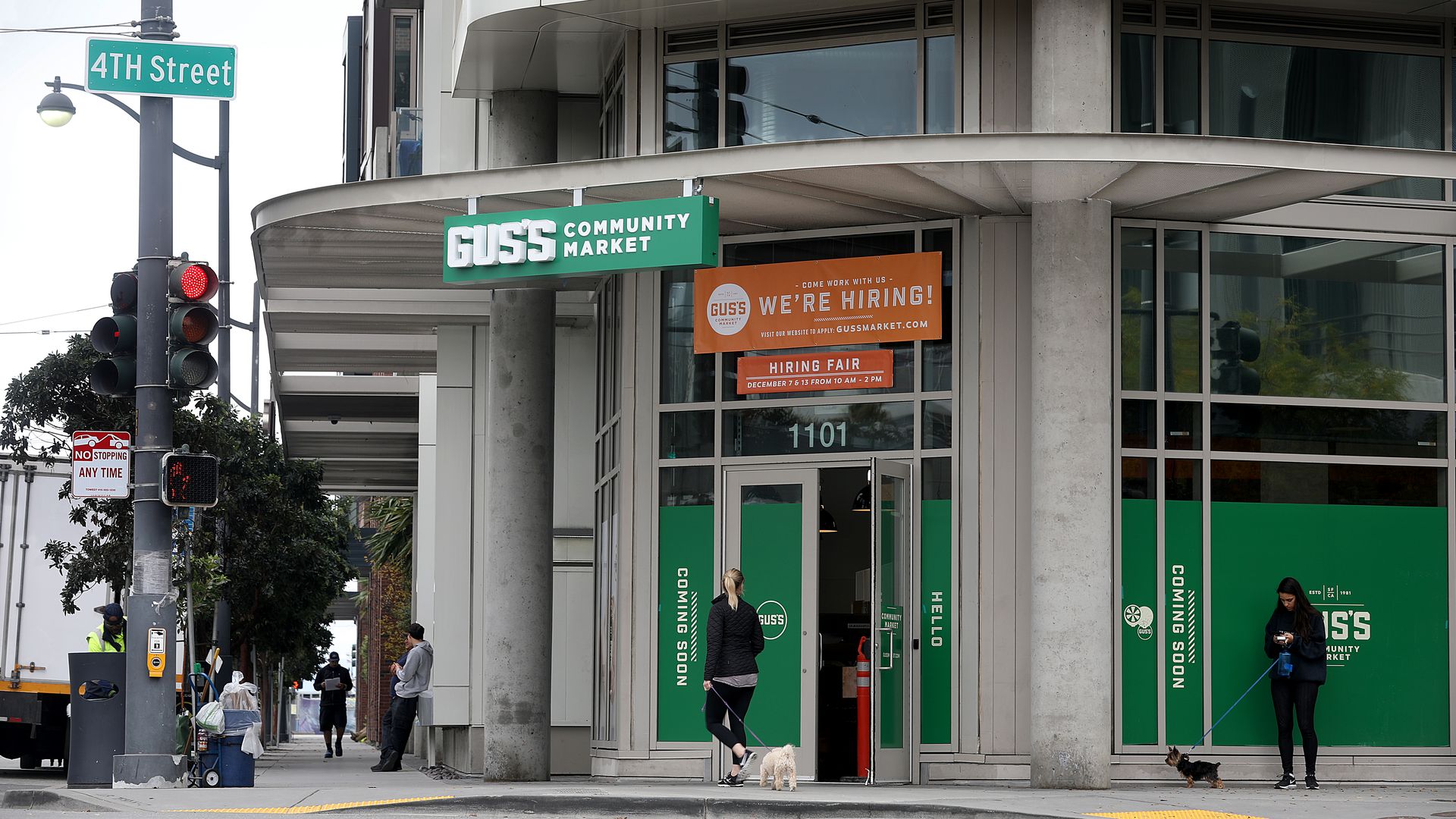 Corner view of a modern building with a green GUSS'S Community Market sign and an orange "WE'RE HIRING!" banner, a 4th Street sign, traffic lights, pedestrians, and small dogs on the sidewalk.