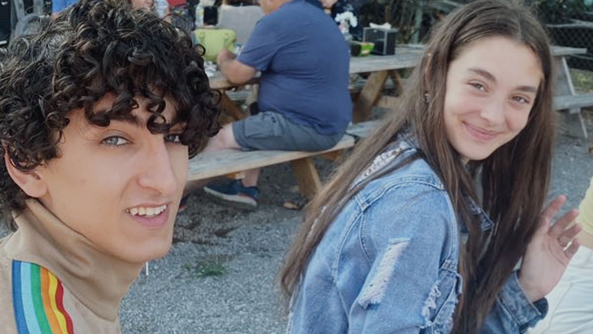 Two teens sit at an outdoor picnic area with wooden benches. A curly-haired boy in a beige shirt with a rainbow stripe looks at the camera; a smiling girl with long hair wears a denim jacket.