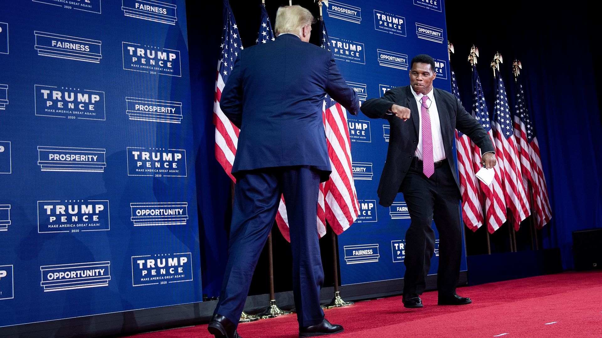 Donald Trump elbow bumps Herschel Walker on a red carpet in front of a wall displaying TRUMP/PENCE signs and a row of American flags.