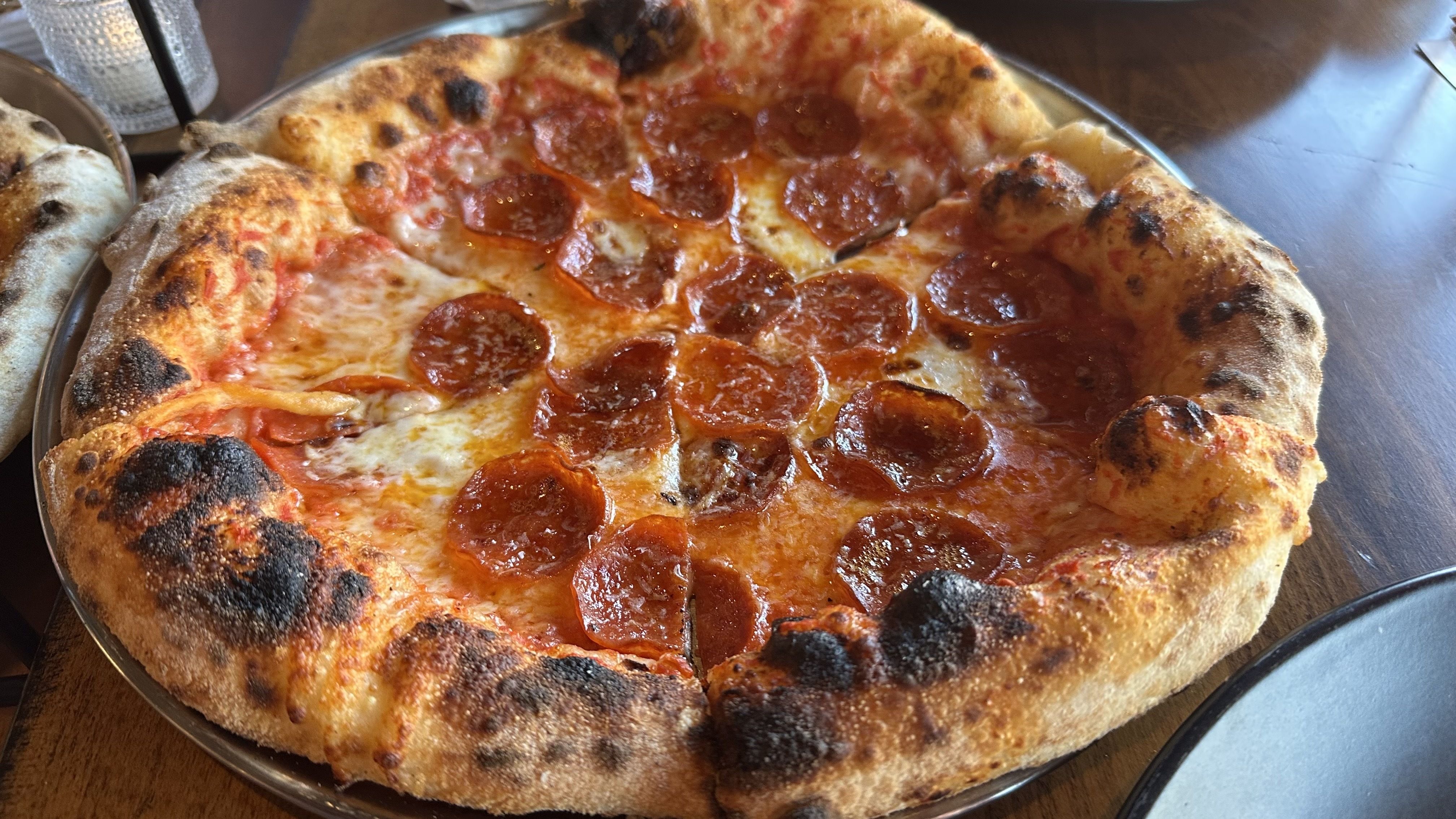 Close-up view of a pepperoni pizza on a metal tray: bubbling mozzarella, red tomato sauce visible around edge, plenty of pepperoni slices, and a blistered, lightly charred crust.
