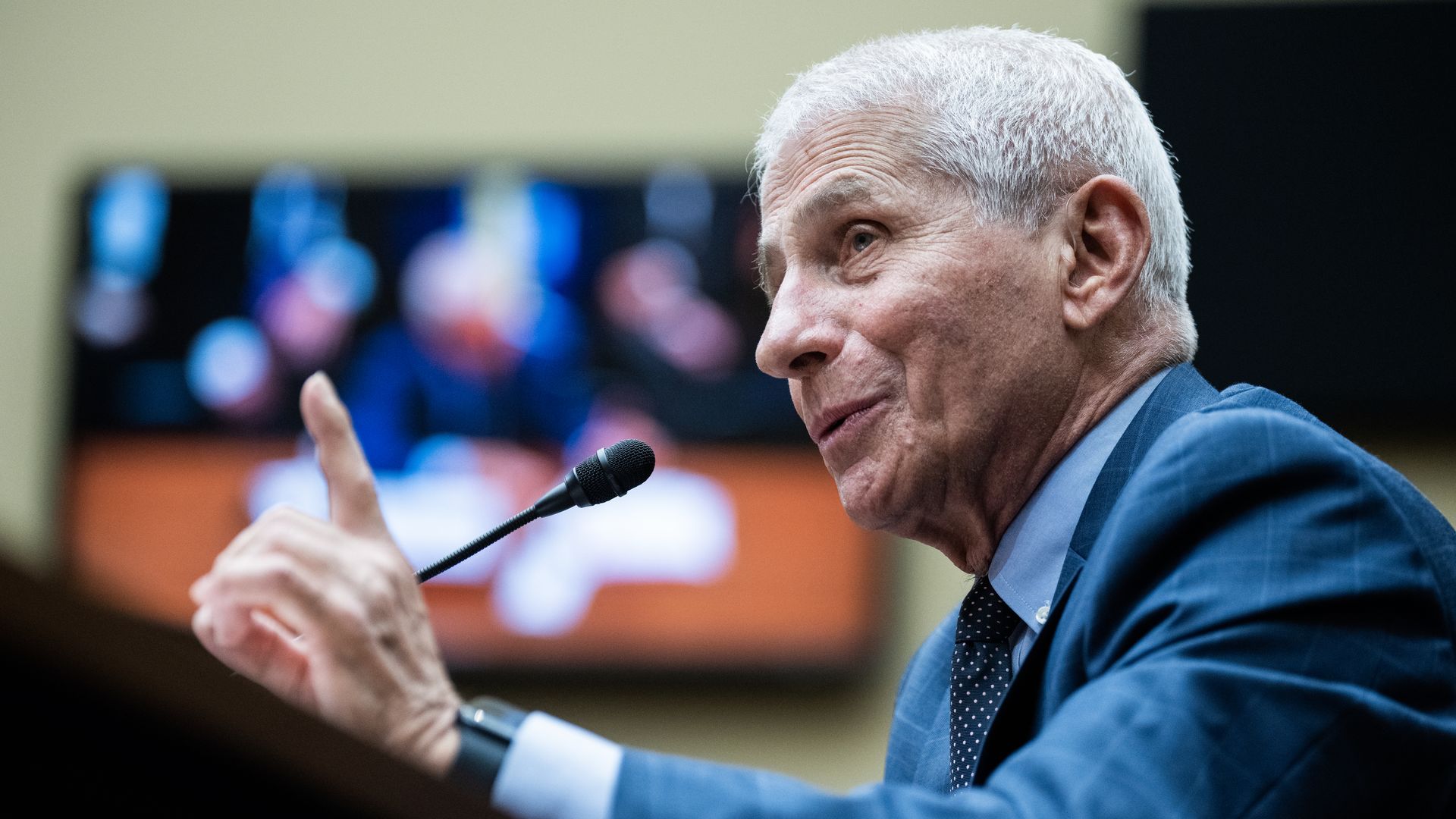 Anthony Fauci speaks into a small microphone. A hand is in front of him with an index finger pointing up. He is wearing a dark suit and light shirt. 