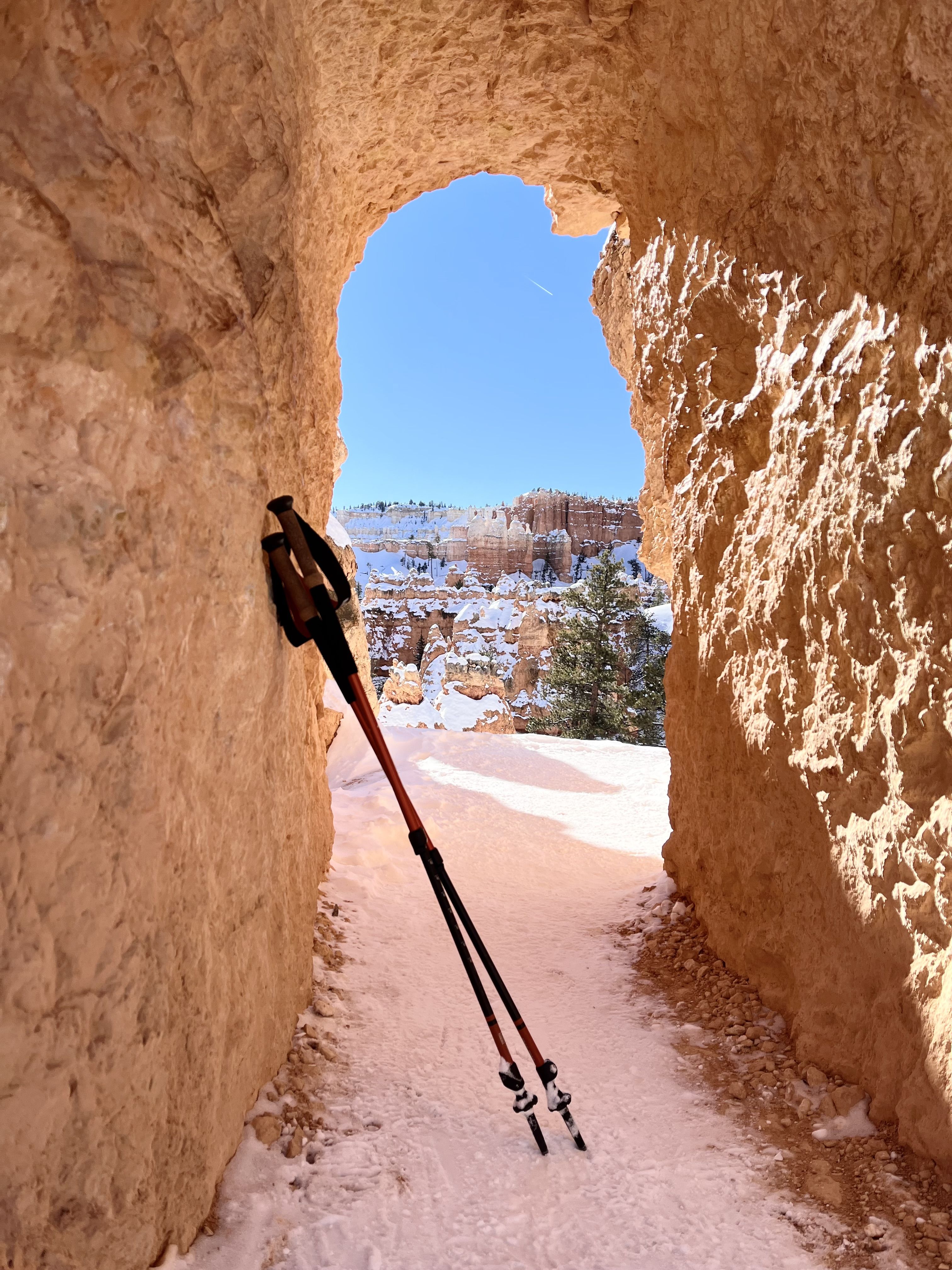 Snowy canyon passage with orange rock walls framing a bright blue sky. A trekking pole leans on the left wall; distant snow-covered cliffs and pine trees dot the opening.
