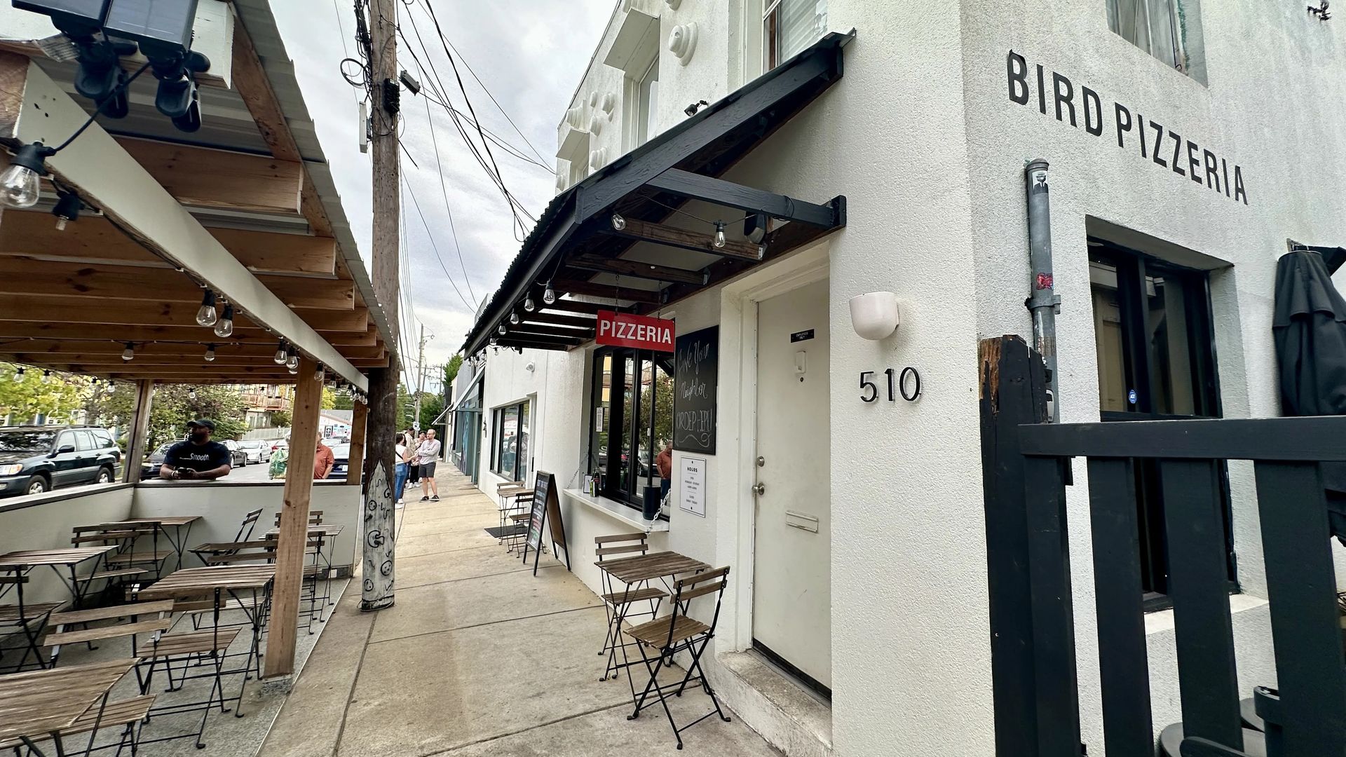 Outdoor seating along a white building labeled BIRD PIZZERIA with a red PIZZERIA sign, string lights, wooden tables, a utility pole, and parked cars on a cloudy day.