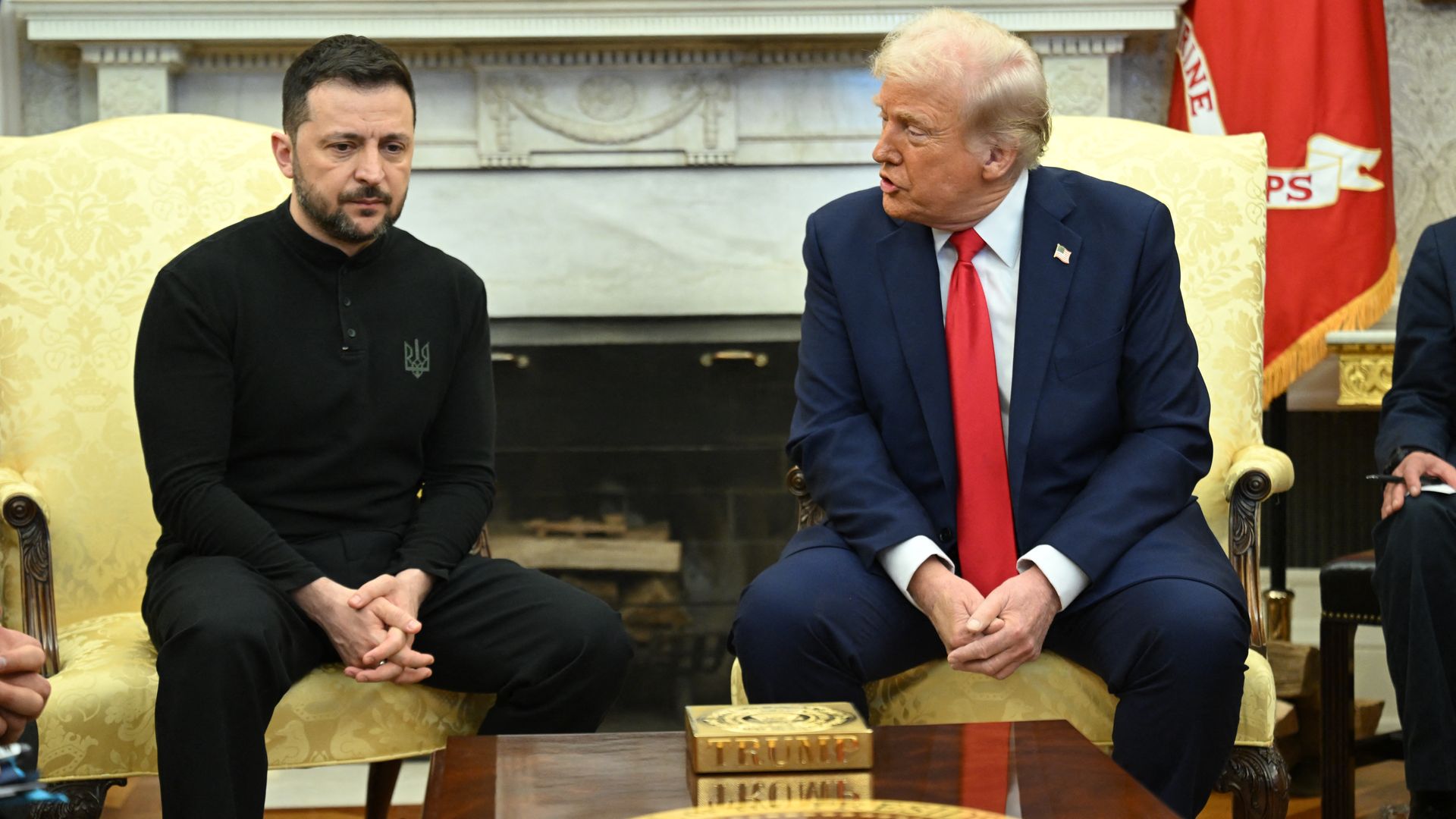 Two men seated in a formal room with ornate decor, one in a black outfit with a serious expression, the other in a dark blue suit and red tie talking and looking at him.