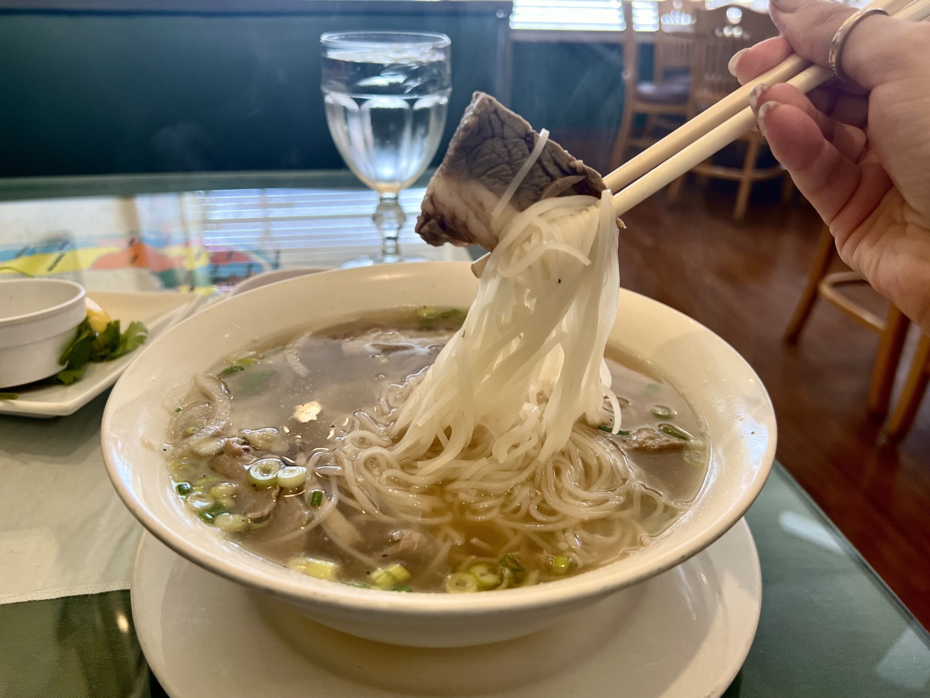 A bowl of Vietnamese pho soup with white rice noodles, slices of beef, and green onions in a clear broth. A hand is holding chopsticks picking up a portion of noodles with a piece of beef. A glass of water and a small plate with garnishes like lemon and cilantro are in the background on the table.