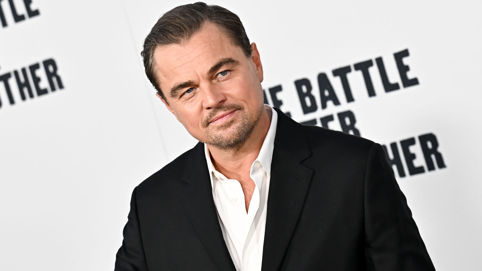 Man with slicked-back hair and light beard wearing a black suit jacket and white shirt standing in front of a white backdrop with partially visible black text.