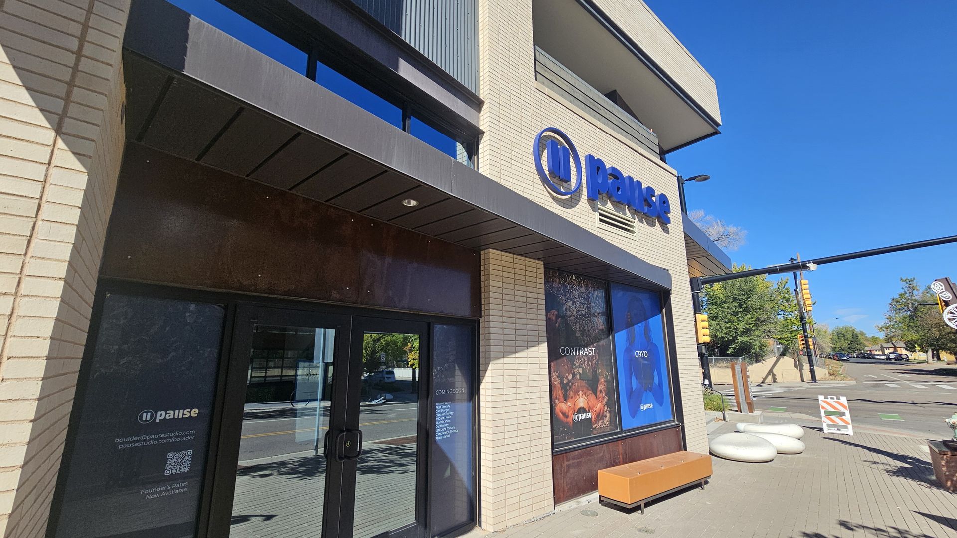 Exterior of a beige brick commercial building with a blue Pause sign on the facade; large glass doors and posters in the windows, blue sky, and a street with traffic lights in the background.