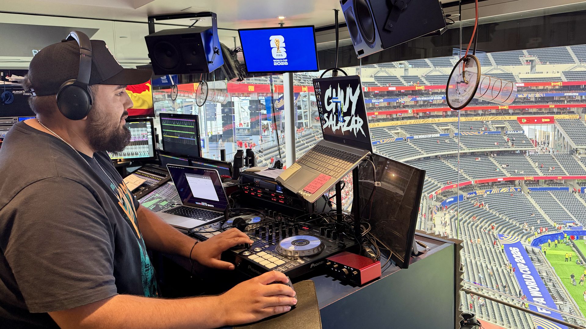 A bearded man wearing a black cap and headphones sits at a broadcast control station inside a stadium booth, surrounded by laptops, screens, and audio gear, with empty stands visible outside.